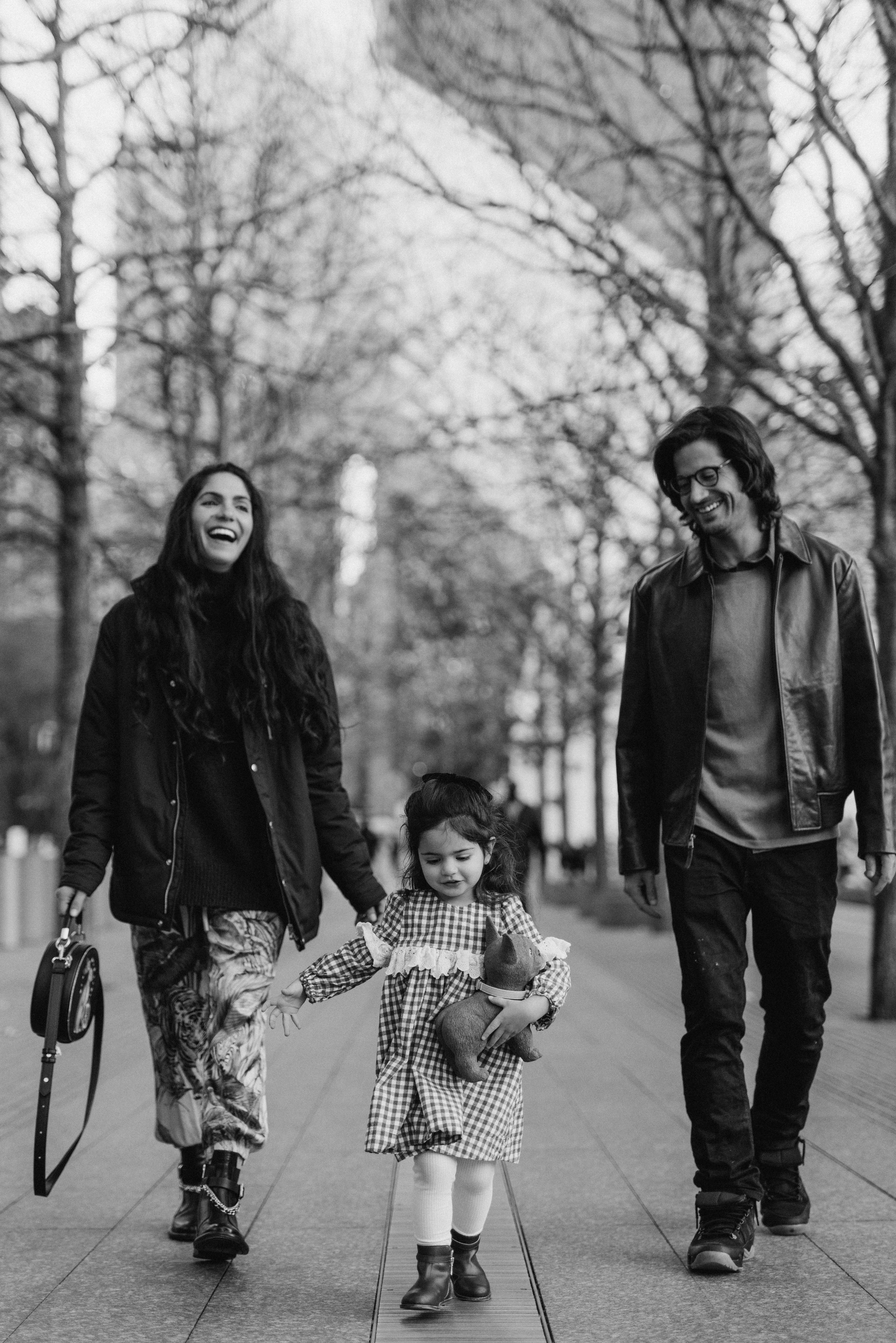 A family portrait in NYC of three, a woman, man, and young girl, walking and smiling outdoors in a park with leafless trees, the girl holding a stuffed animal, by Maison Mancel.