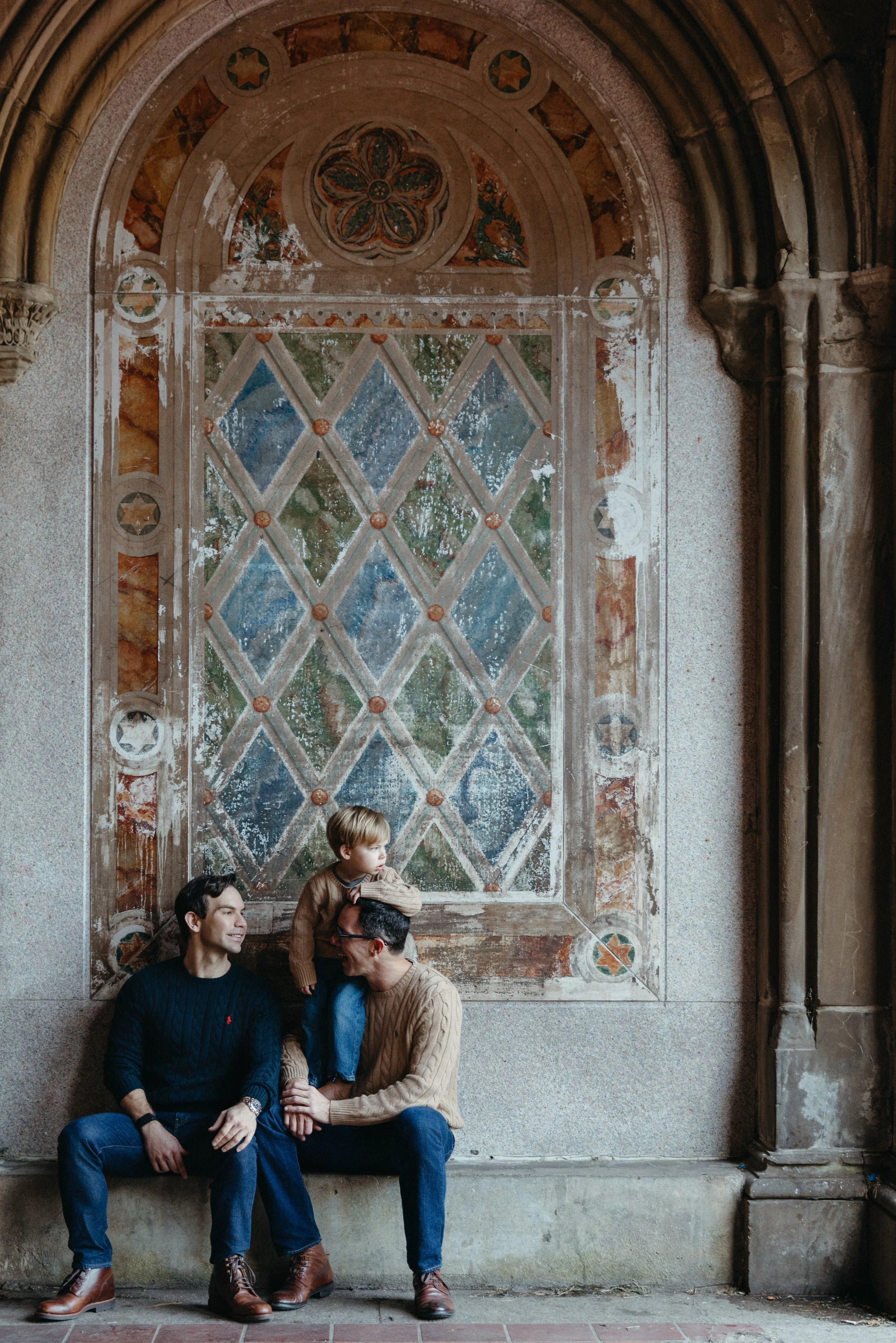 A family photograph of two men and a young son sitting on a bench in front of an ornate stained glass window in Central Park, by Maison Mancel.