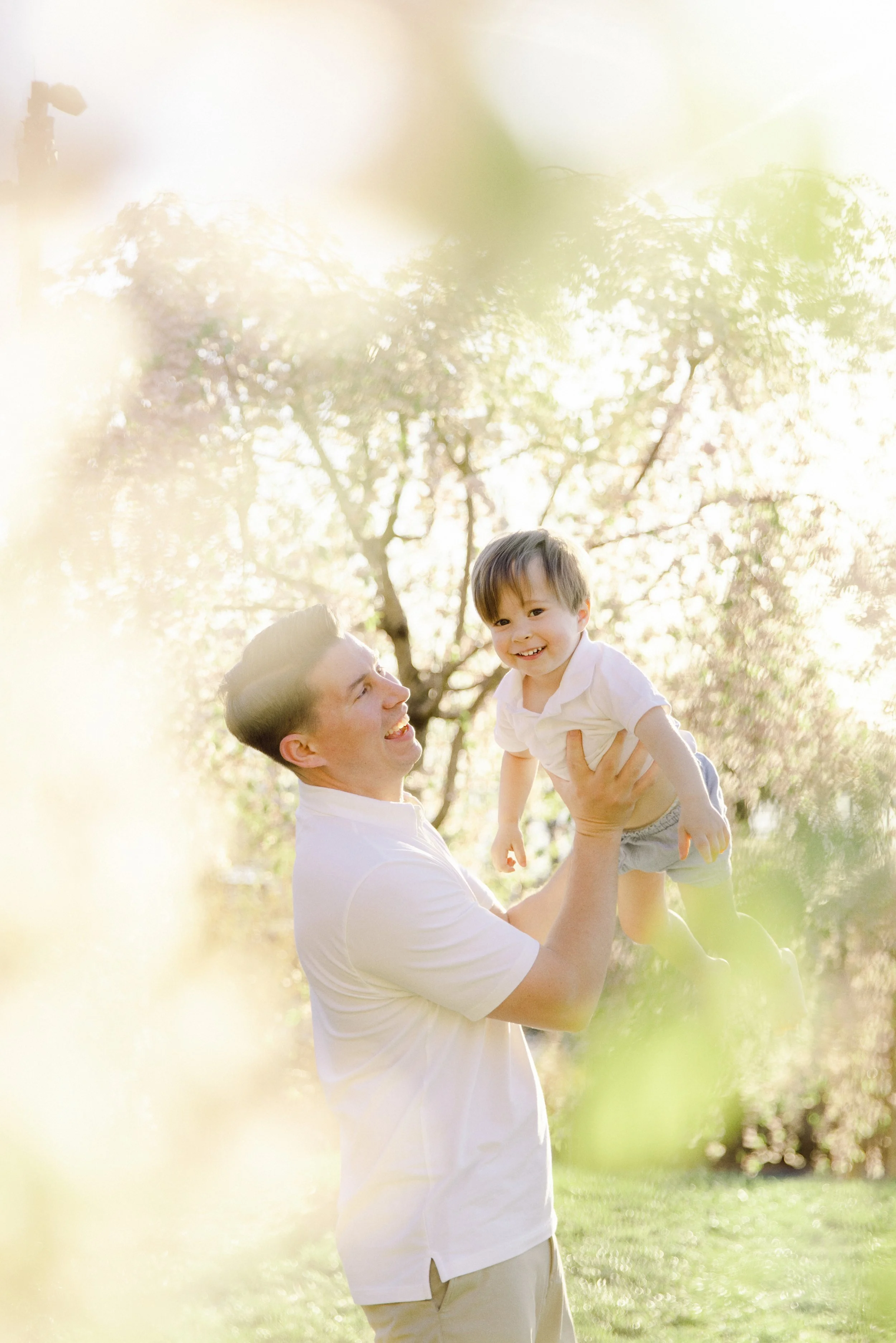 A portrait of a father playing with a young son outdoors during daytime, surrounded by blossoming trees with sunlight filtering through the branches in Brooklyn, by Maison Mancel.