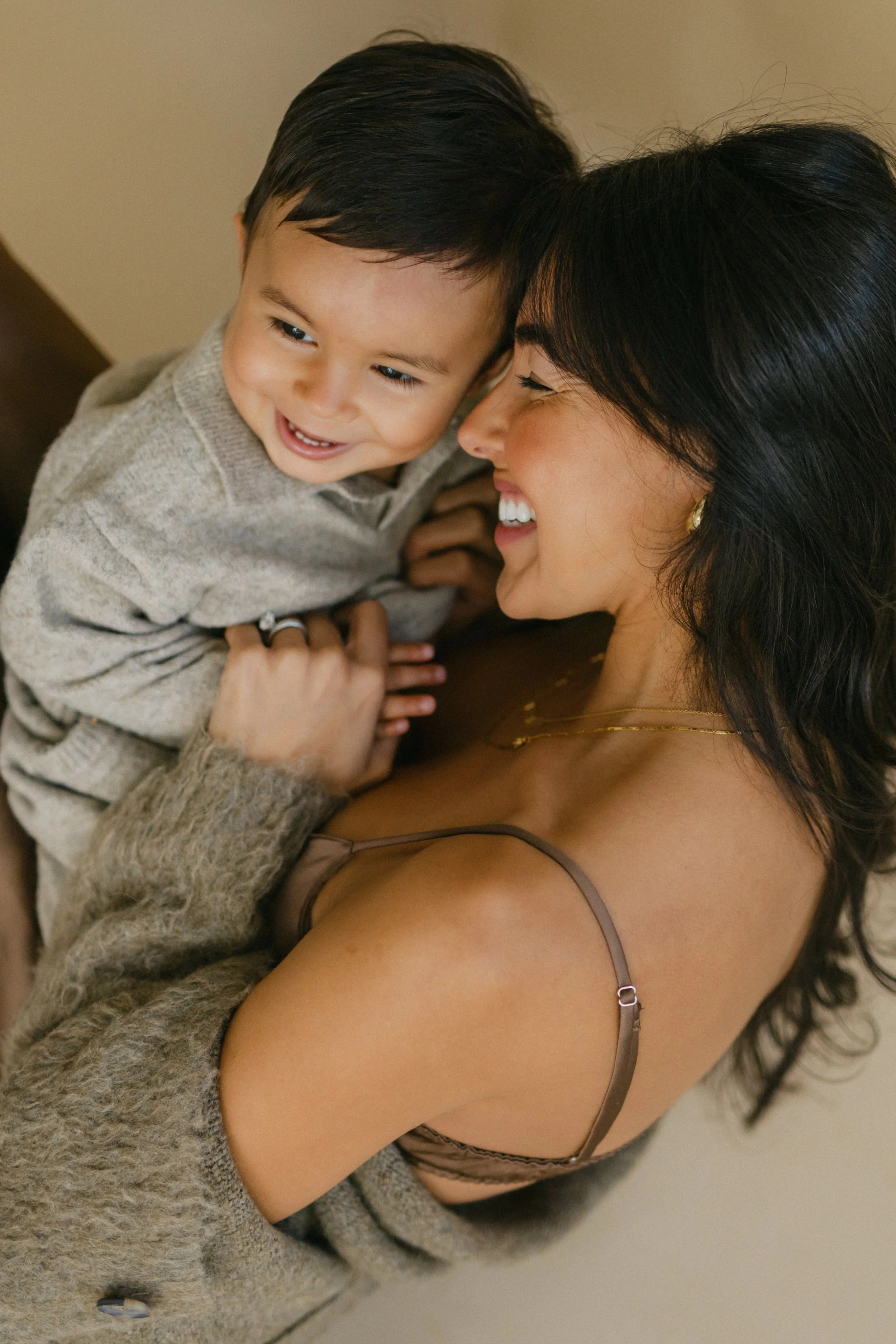 An in-studio family portrait of a mother with dark hair smiling and holding a young boy close, both smiling and looking at each other - Maison Mancel.