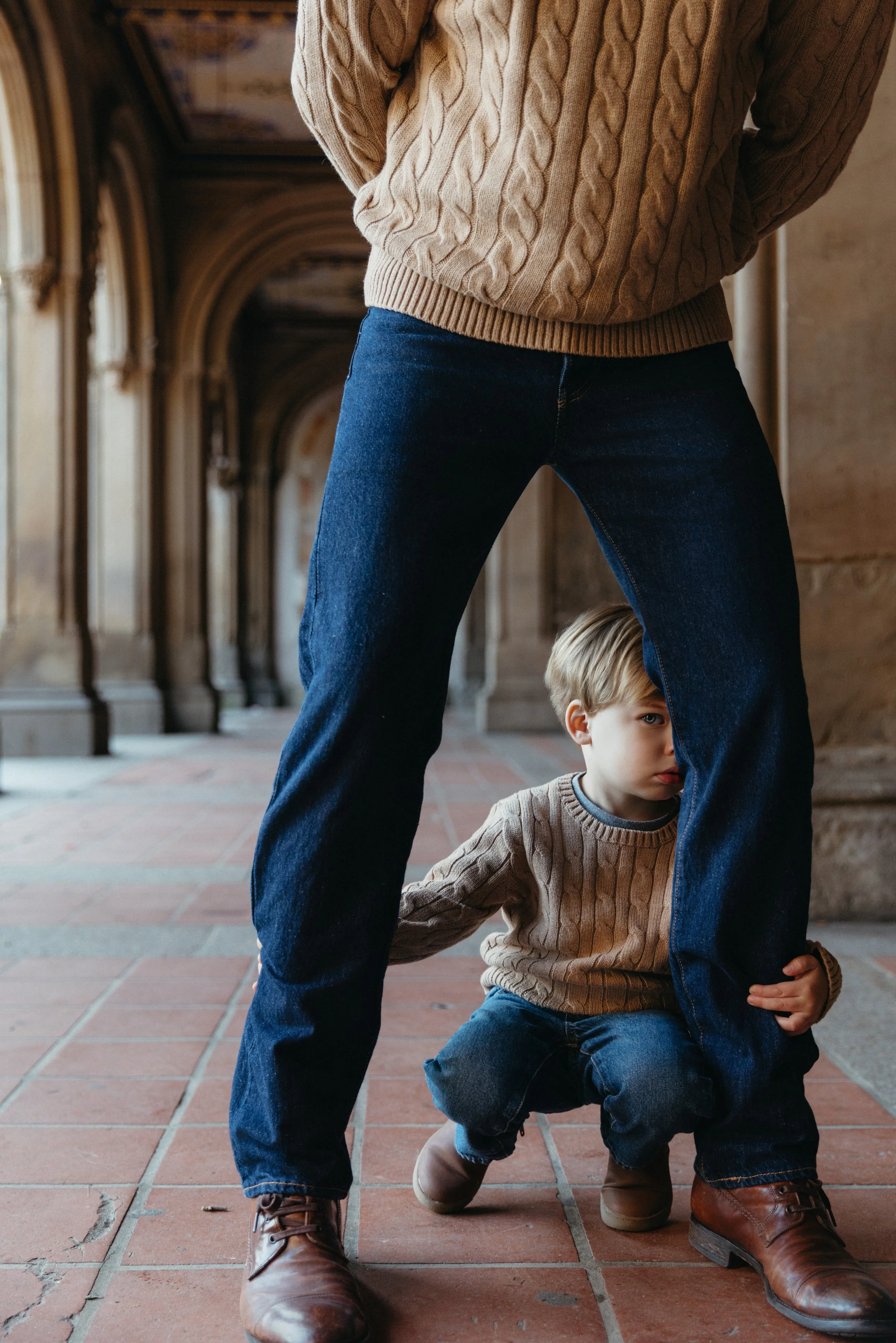 A portrait of person wearing a beige cable-knit sweater and blue jeans standing in a corridor, with a young boy crouching underneath their legs, holding onto the person's leg and looking to the side, taken in Central Park by Maison Mancel.