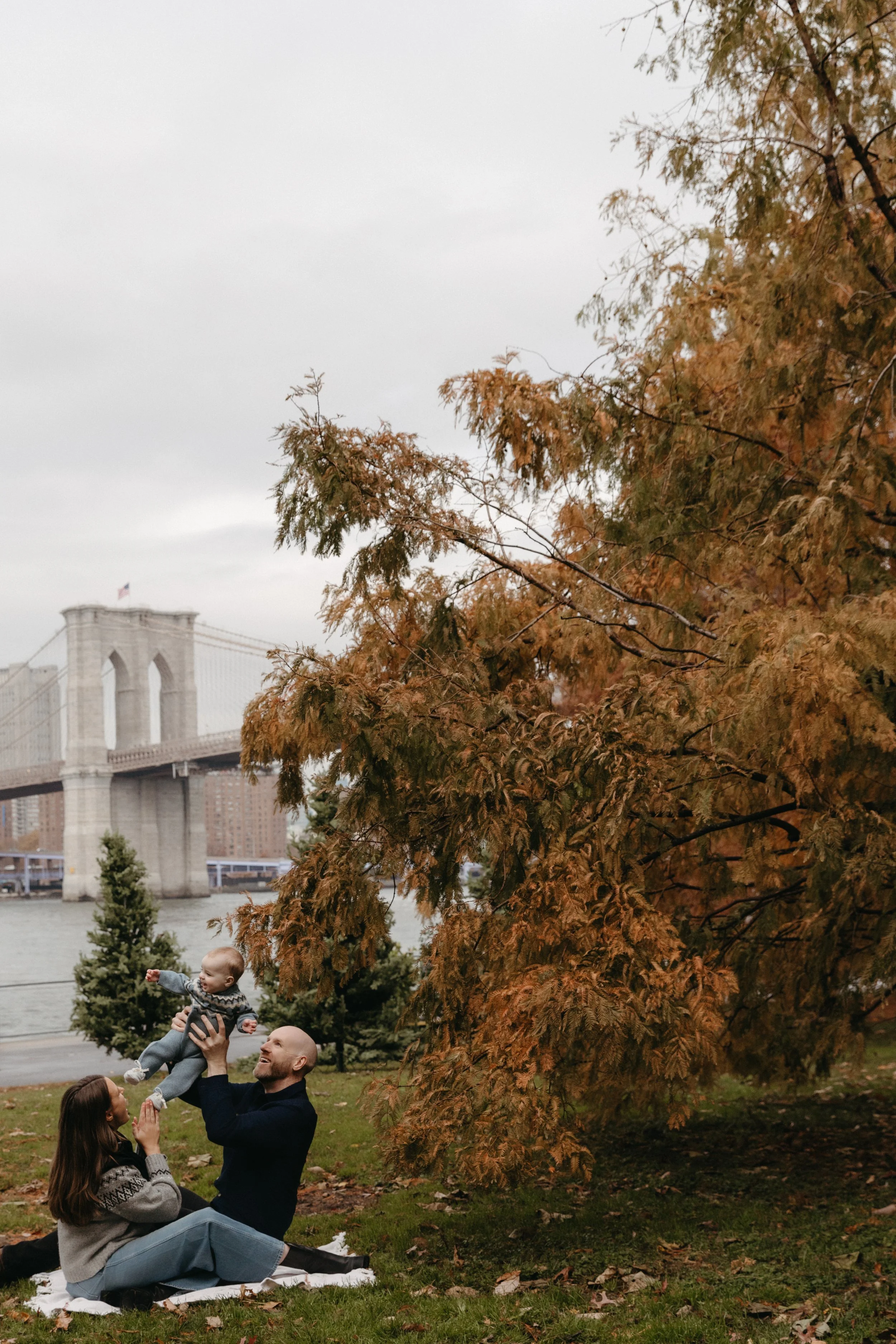 A family of three enjoying time outdoors in Brooklyn Bridge park with an autumn tree, the Brooklyn Bridge, and city buildings in the background, photographed by Maison Mancel.