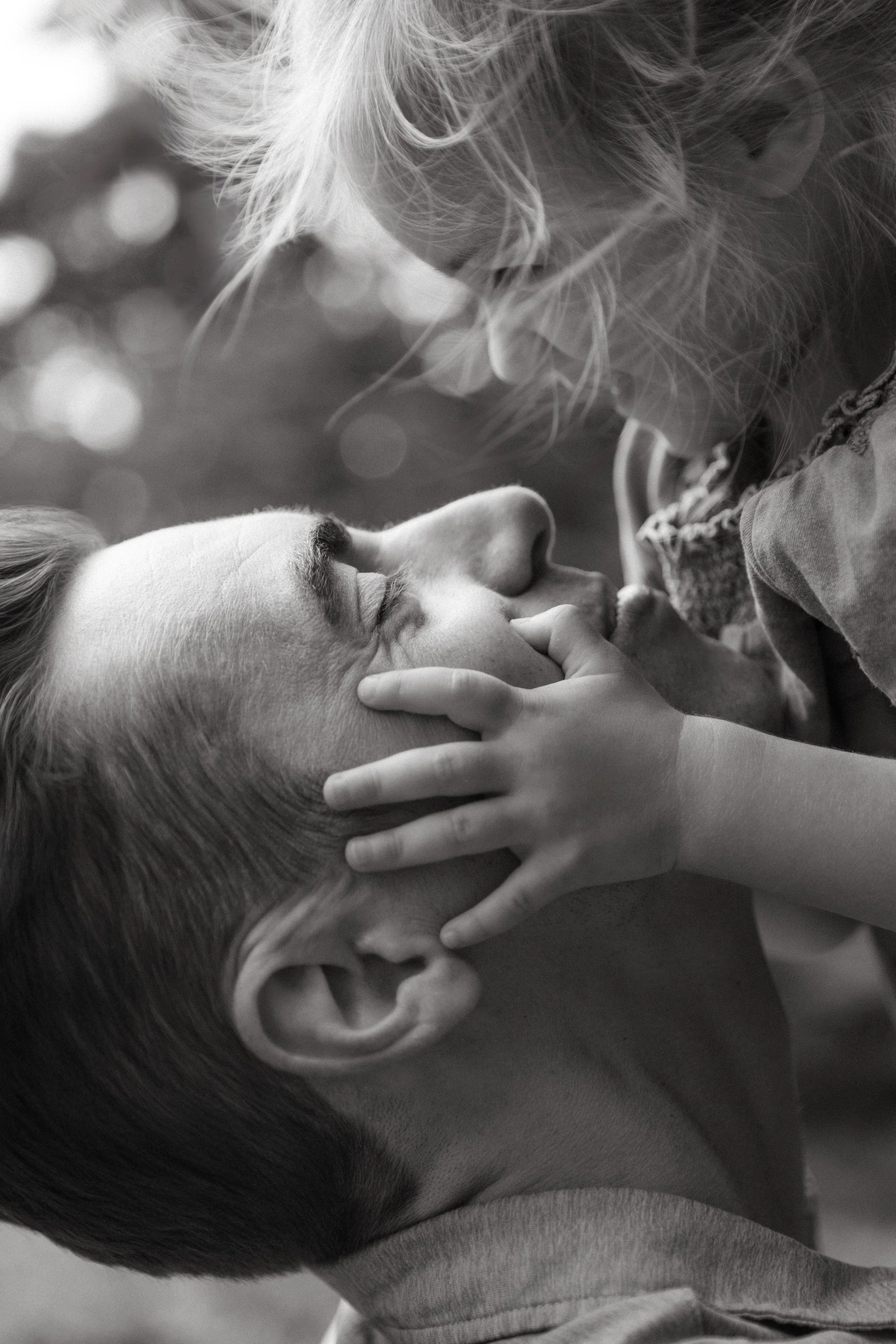 A black and white family photograph of a young girl and her father touching their noses together, with the girl holding the father's face gently, in Central Park, by Maison Mancel.