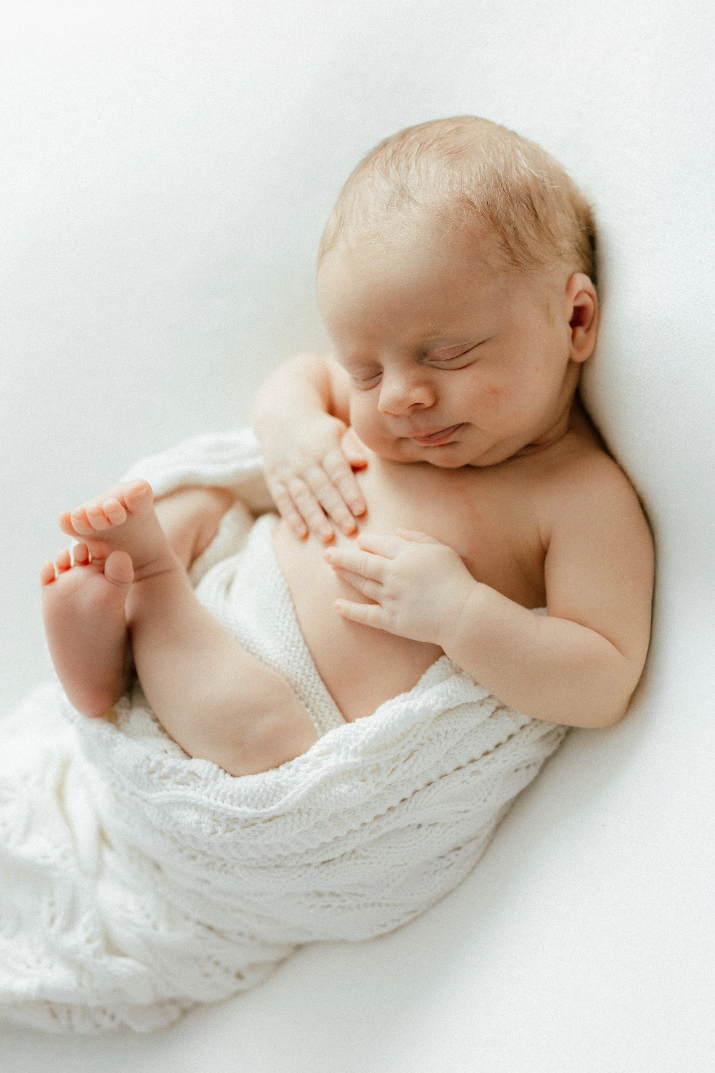 A sleeping newborn baby wrapped in a white knitted blanket, lying on a white surface, photographed by Maison Mancel in NYC.