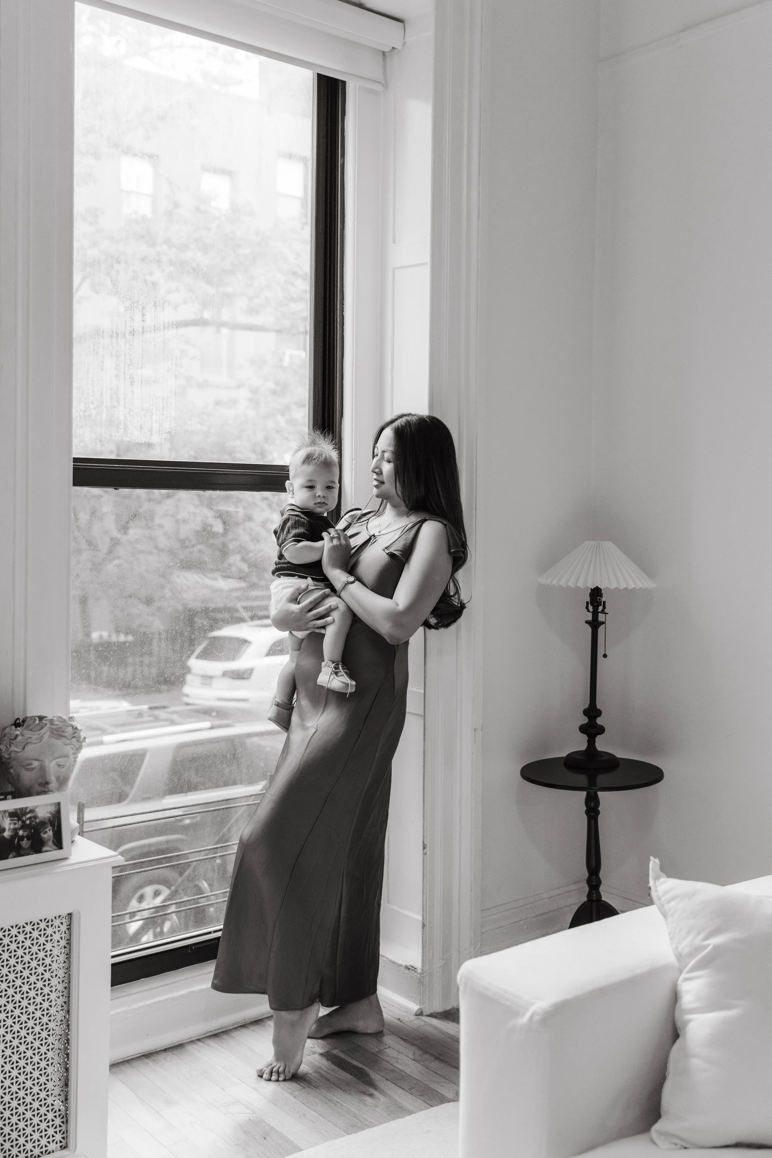 An in-home lifestyle family portrait of a mother holding a young son near a window in a home, with a lamp and framed photo on a table in the background, taken in Brooklyn by Maison Mancel.