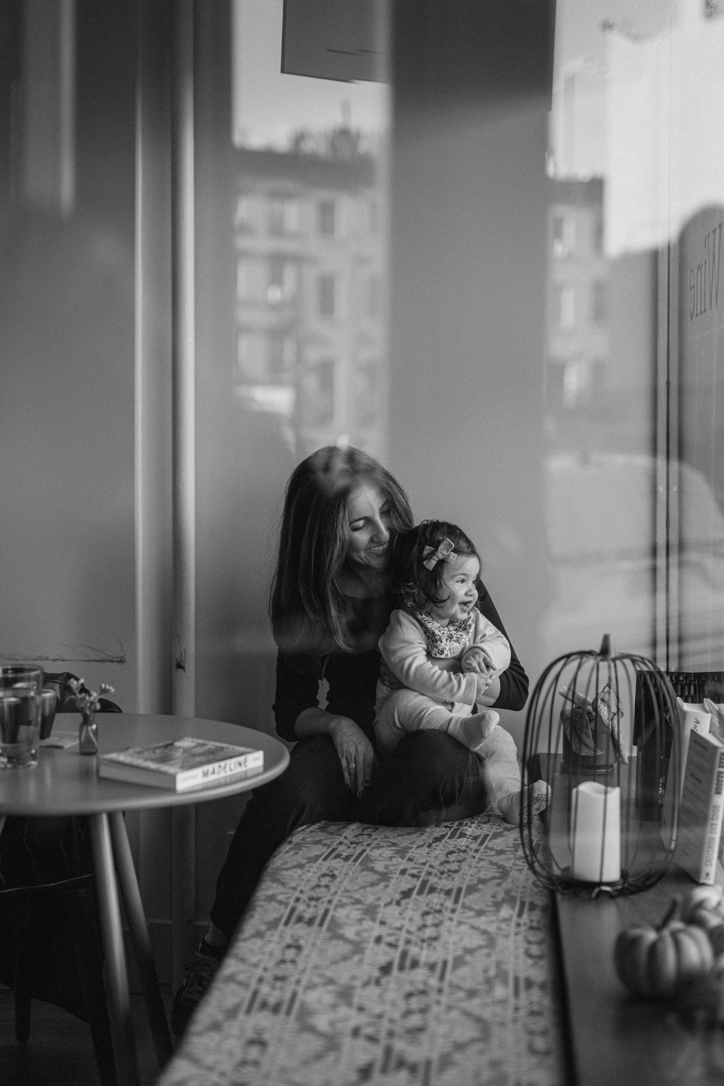 A family portrait of a mother  and a young girl sitting together, looking out a window with reflections of buildings. The girl is smiling, and the scene is in black and white, taken in Carroll Gardens, Brooklyn, by Maison Mancel. 