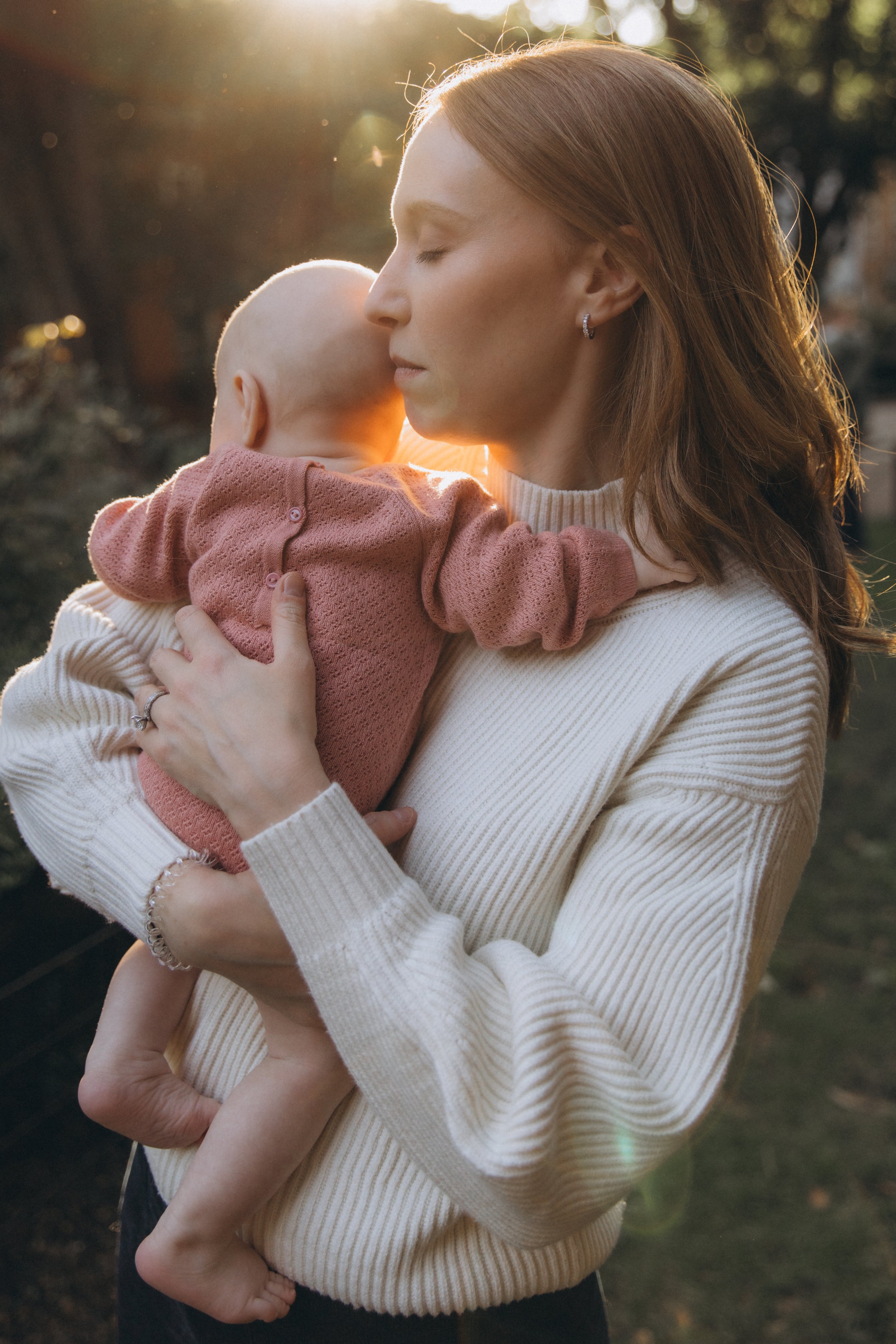 A family portrait of a mother with light skin and red hair holding a sleeping baby with light skin and a bald head, outdoors during sunset, with sunlight glowing behind them - Maison Mancel.