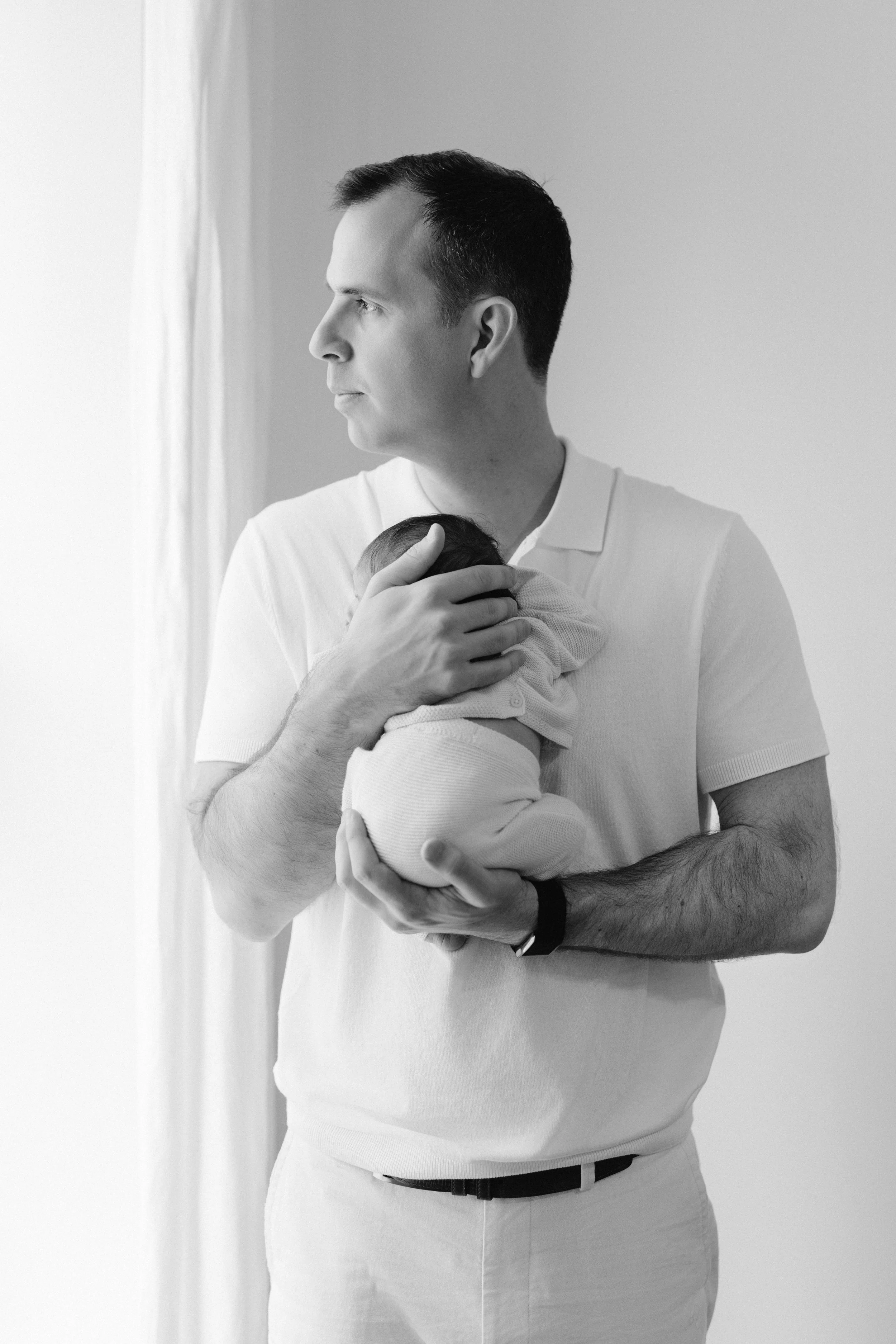A father holding a newborn baby near a window, with a contemplative expression, in black and white, photographed by Maison Mancel in-hom in Manhattan.