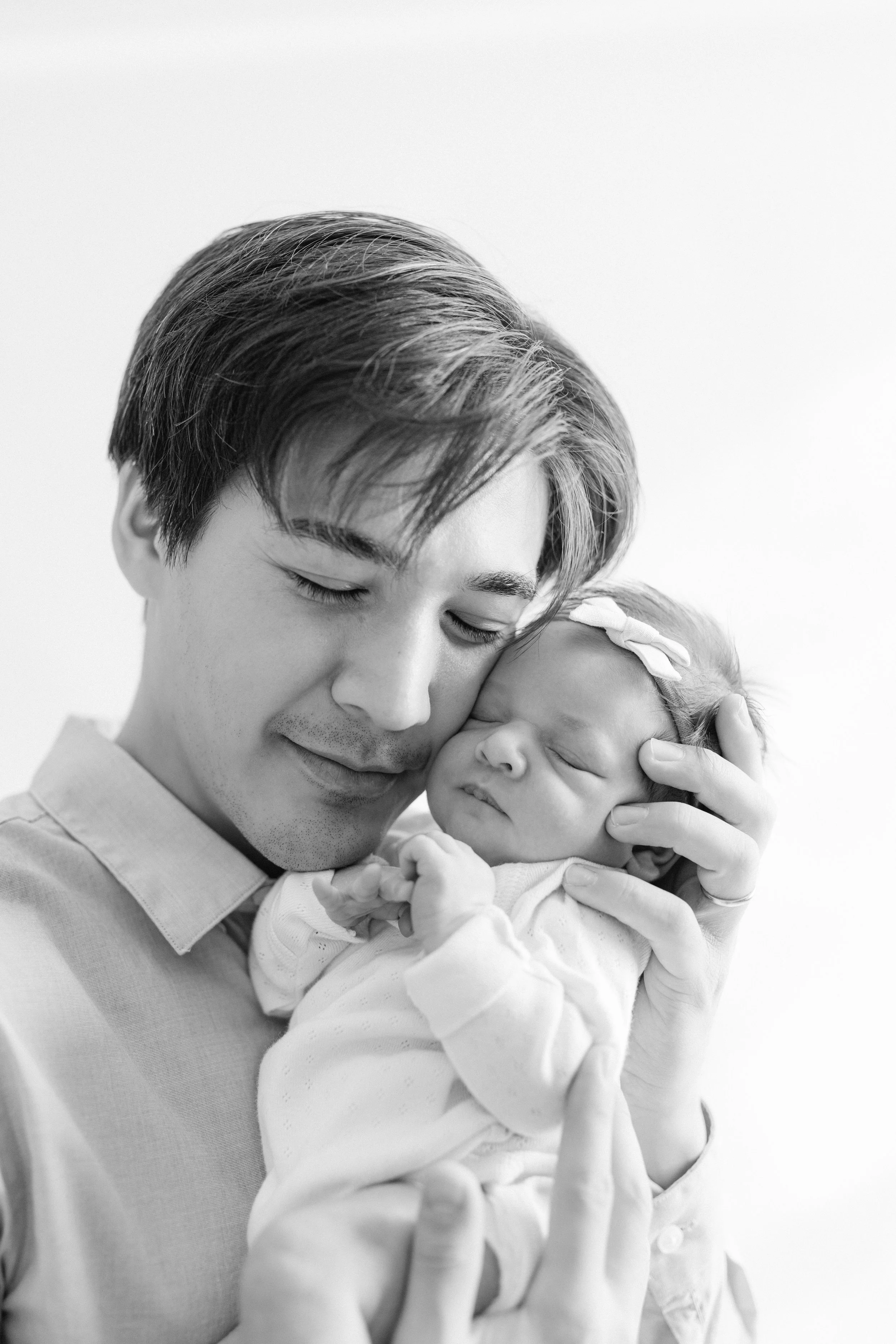 Black and white portrait of a young father holding a sleeping newborn baby close, with their foreheads touching, both with peaceful expressions., Photographed by Maison Mancel in-studio in NYC.