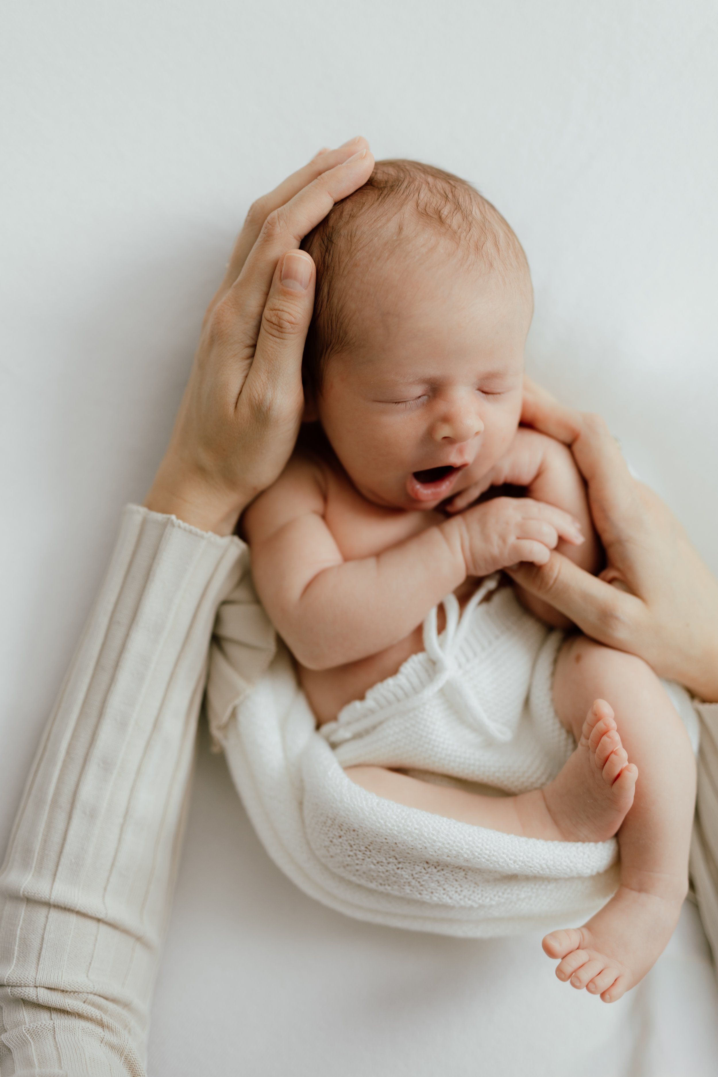 In-studio portrait of a newborn baby boy sleeping on a white blanket with an adult's hand gently cradling his head, photographed by Maison Mancel in NYC.