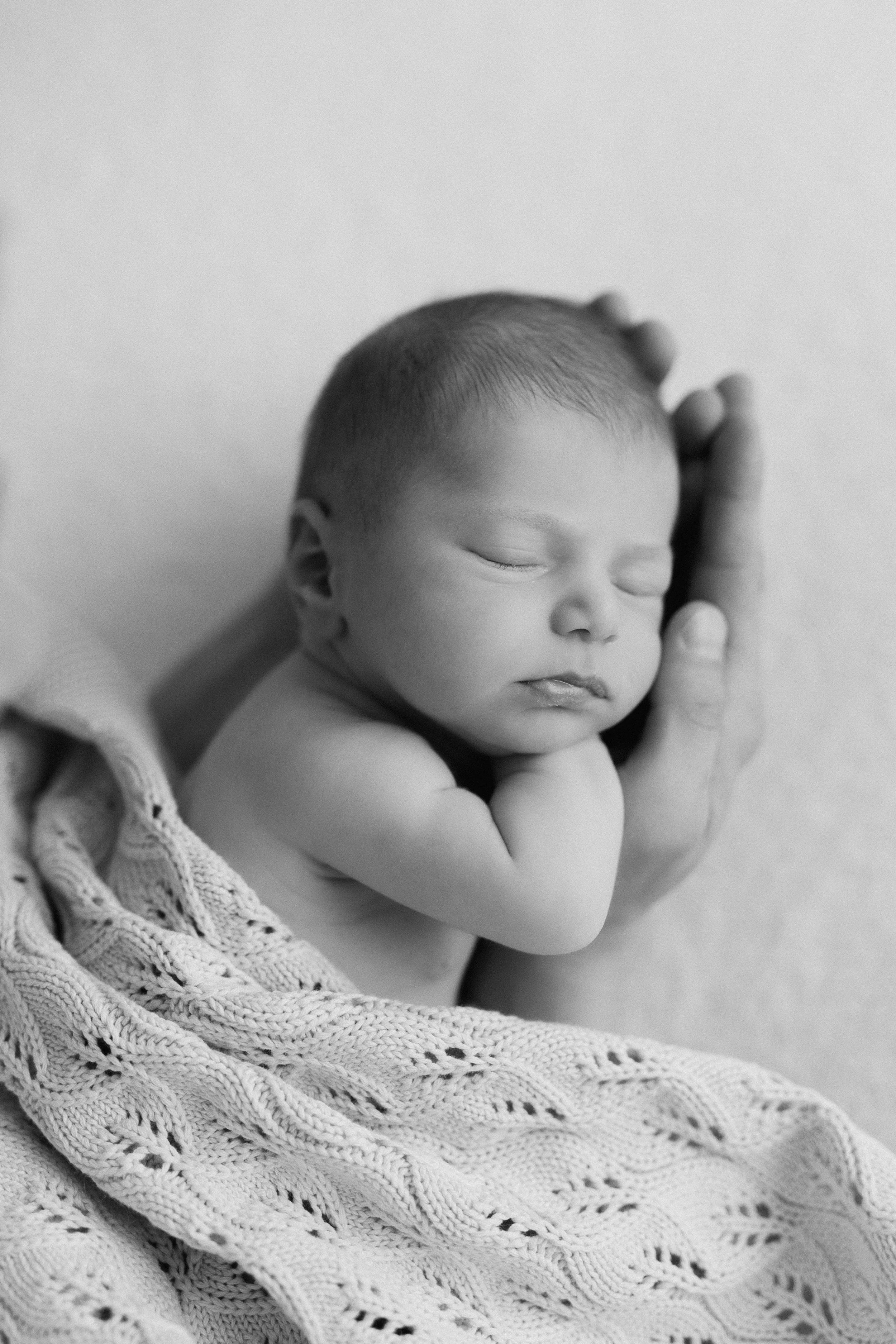 Black and white photo of a sleeping newborn baby with hands under chin, wrapped in a knitted blanket, supported by an adult hand taken in studio by Maison Mancel in NYC.