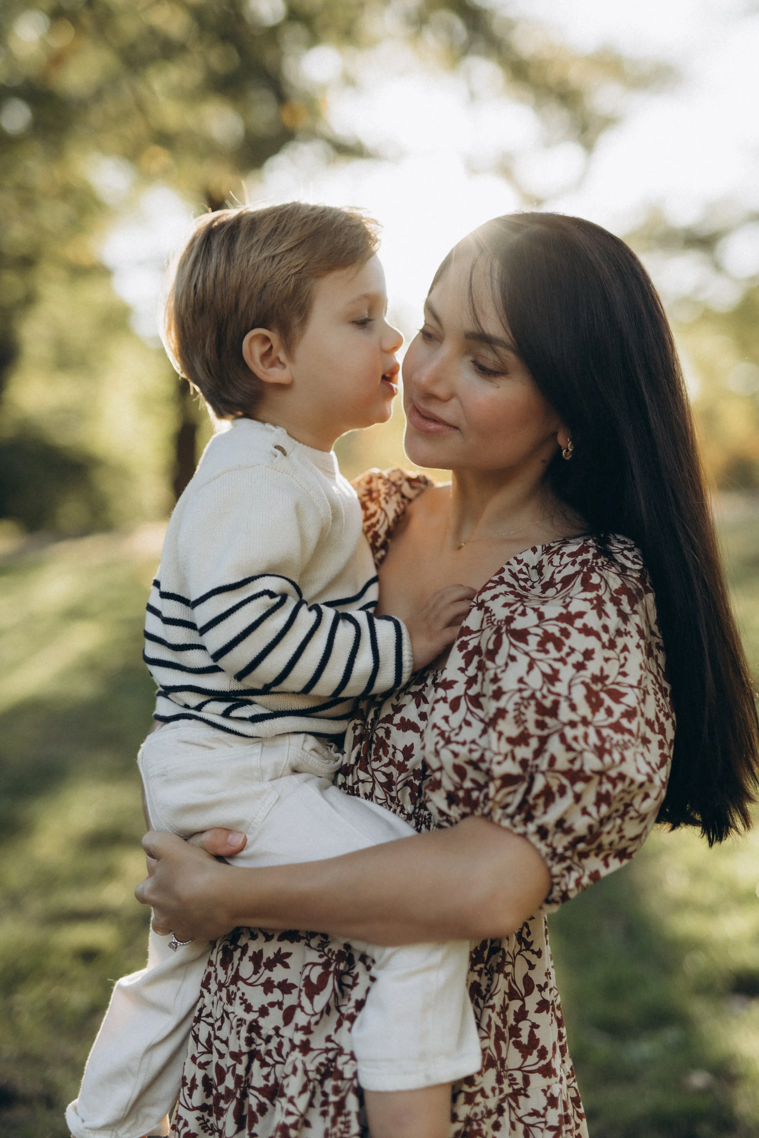 A family portrait in Central Park of a mother holding a young boy in a park with trees in the background, sunlight filtering through the leaves, photographed by Maison Mancel in NYC.