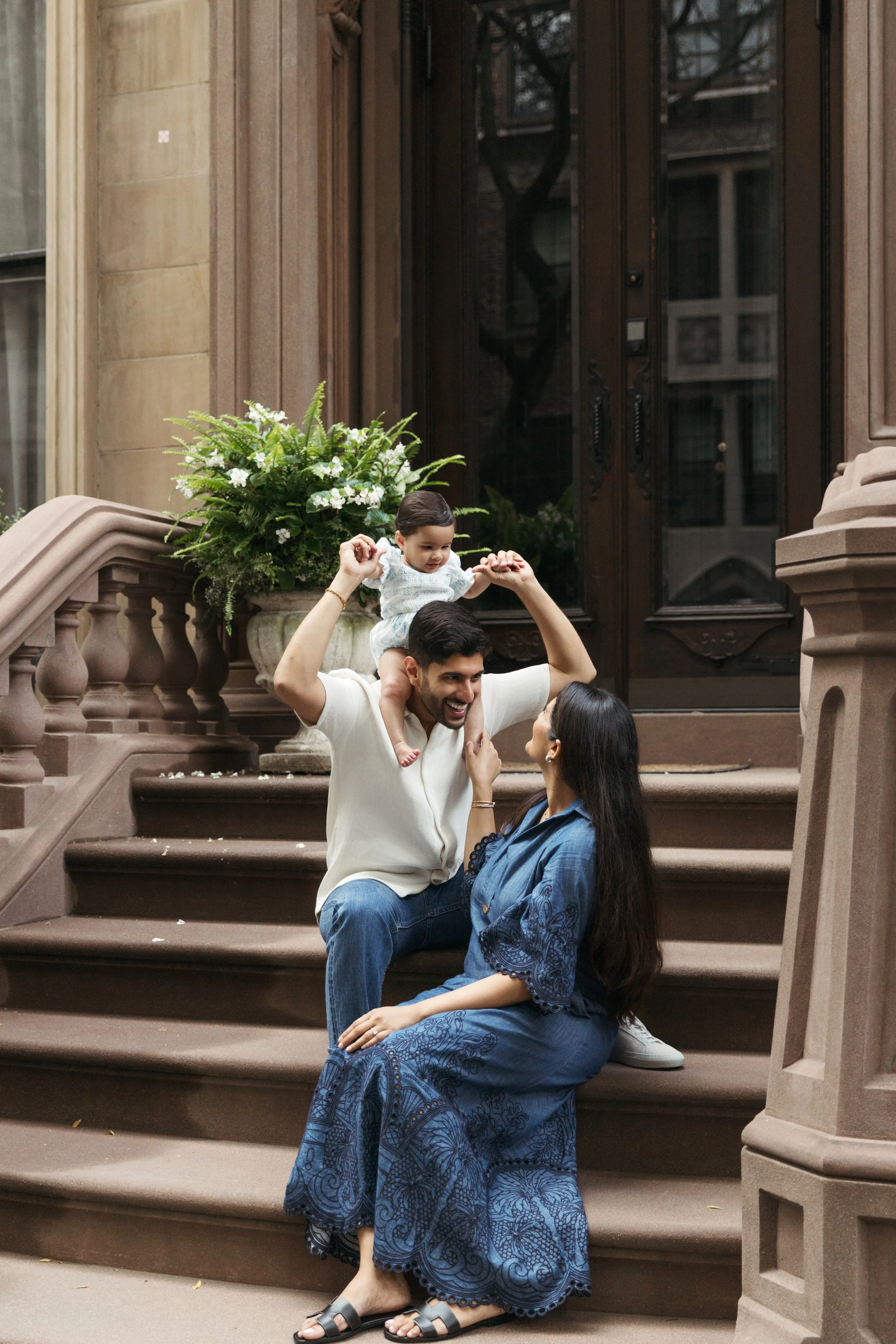 NYC portrait of a family of three sitting on brownstone stairs outside a building. The father, with dark hair and beard, holds the baby on his shoulders. The mother, with long dark hair, sits on the stairs looking up at them. Taken by Maison Mancel.