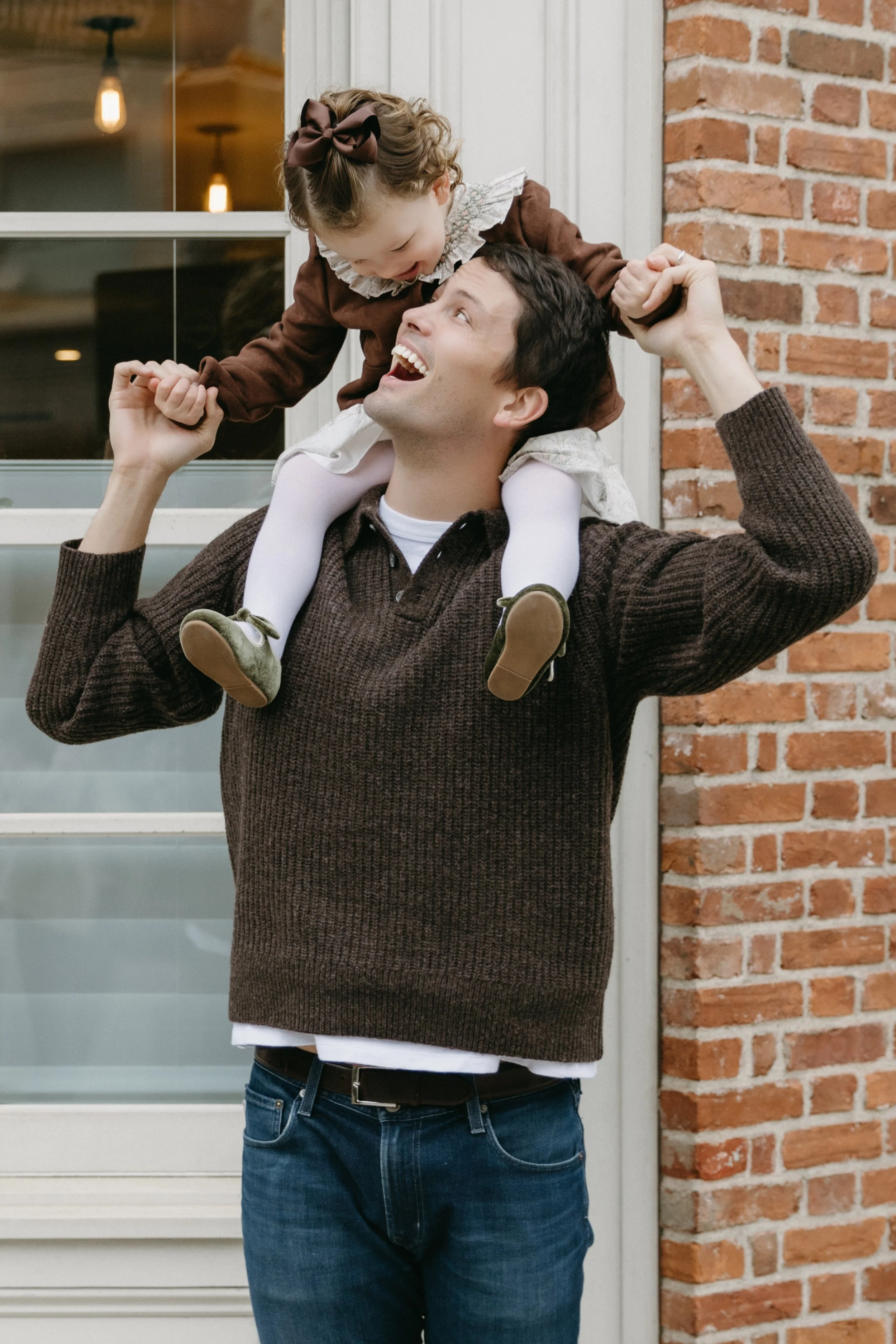 A lifestyle family portrait of a father holding a young girl on his shoulders outside a brick house, both smiling and looking happy, in TriBeca, photographed by Maison Mancel.