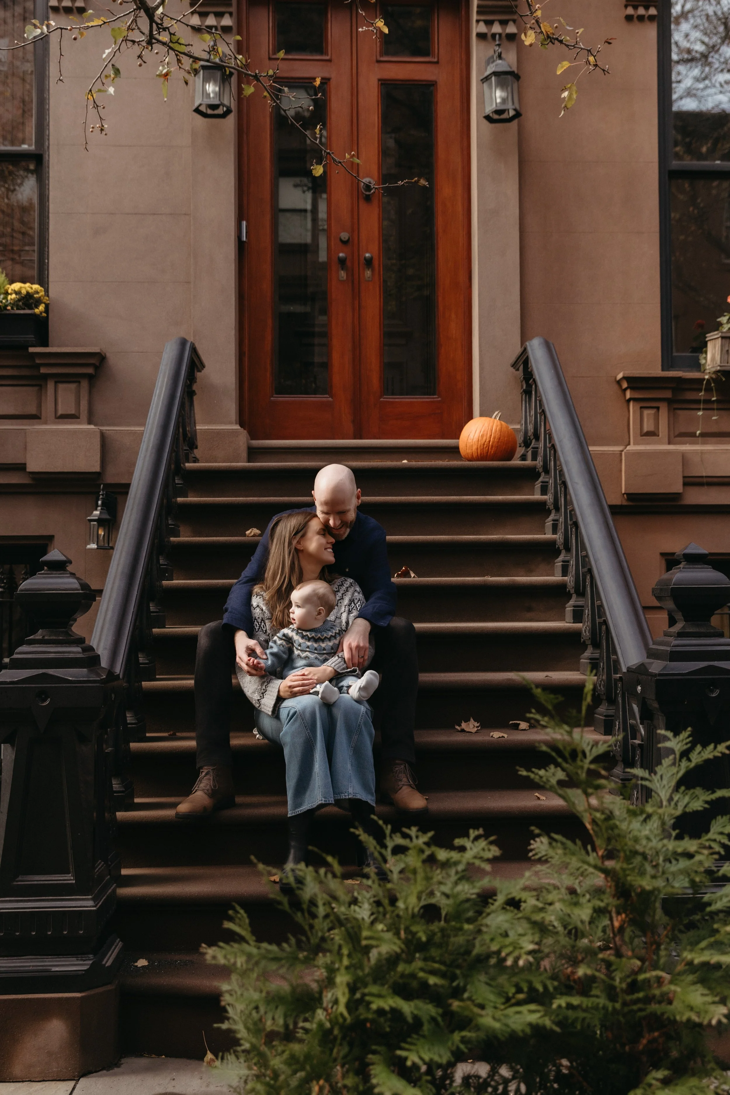 A portrait of a family of three sits on front steps of a Brooklyn brownstone. The parents are laughing and cuddling their child, who is sitting on the mother's lap. The scene is autumnal with fallen leaves and a tree branch with leaves overhead. 