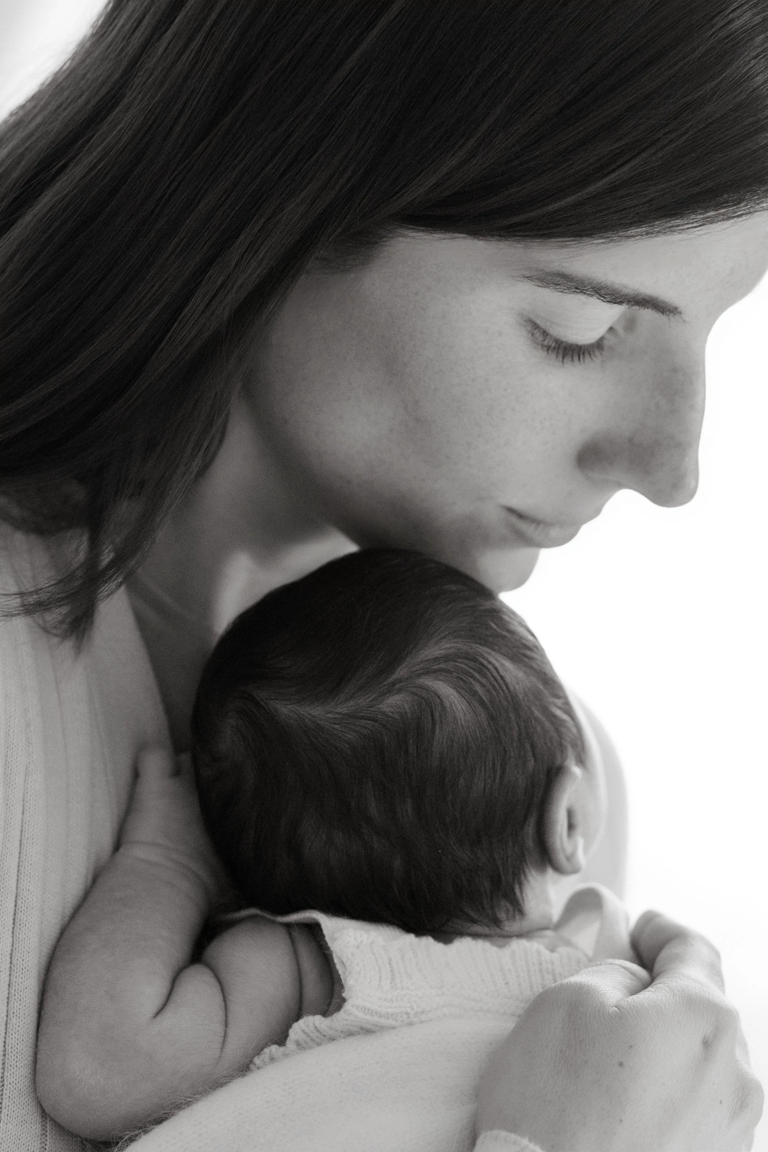 Close-up of a mother with long hair holding a sleeping newborn with dark hair, both in a tender moment, photographed by Maison Mancel in-stdio in NYC.