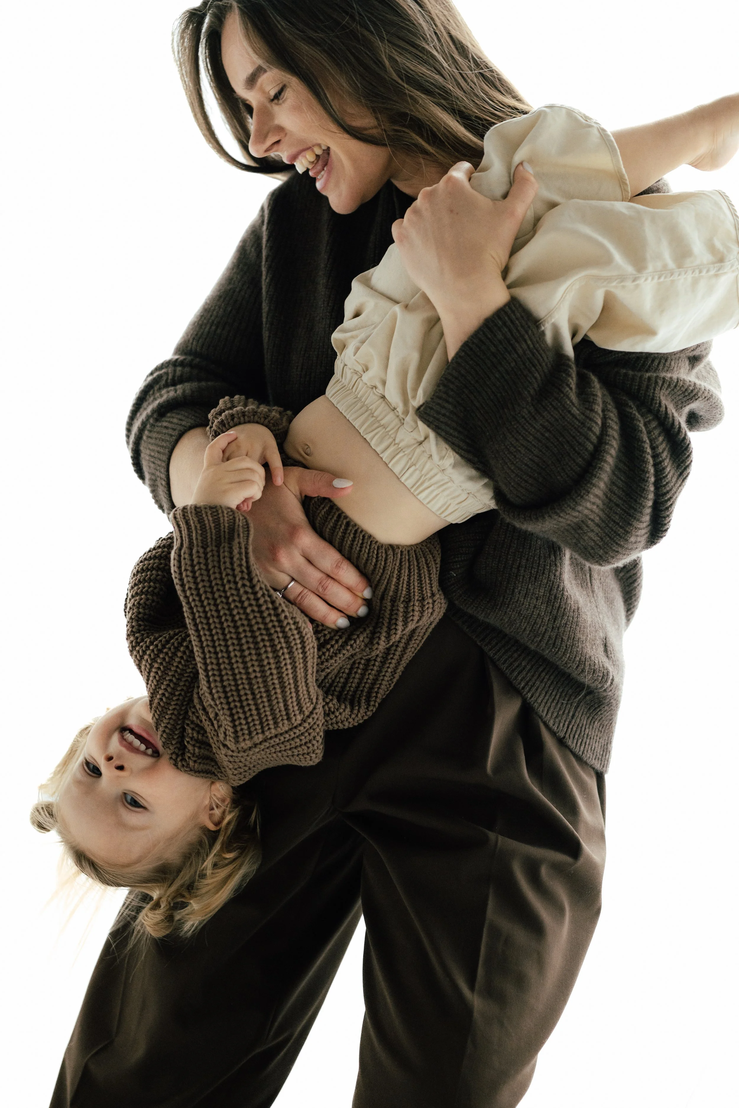 NYC family photograph of a mother and a young girl are playfully holding each other, with the mother smiling at the girl who is hanging upside down, both dressed in cozy, warm clothing, taken in-studio in Brooklyn by Maison Mancel.