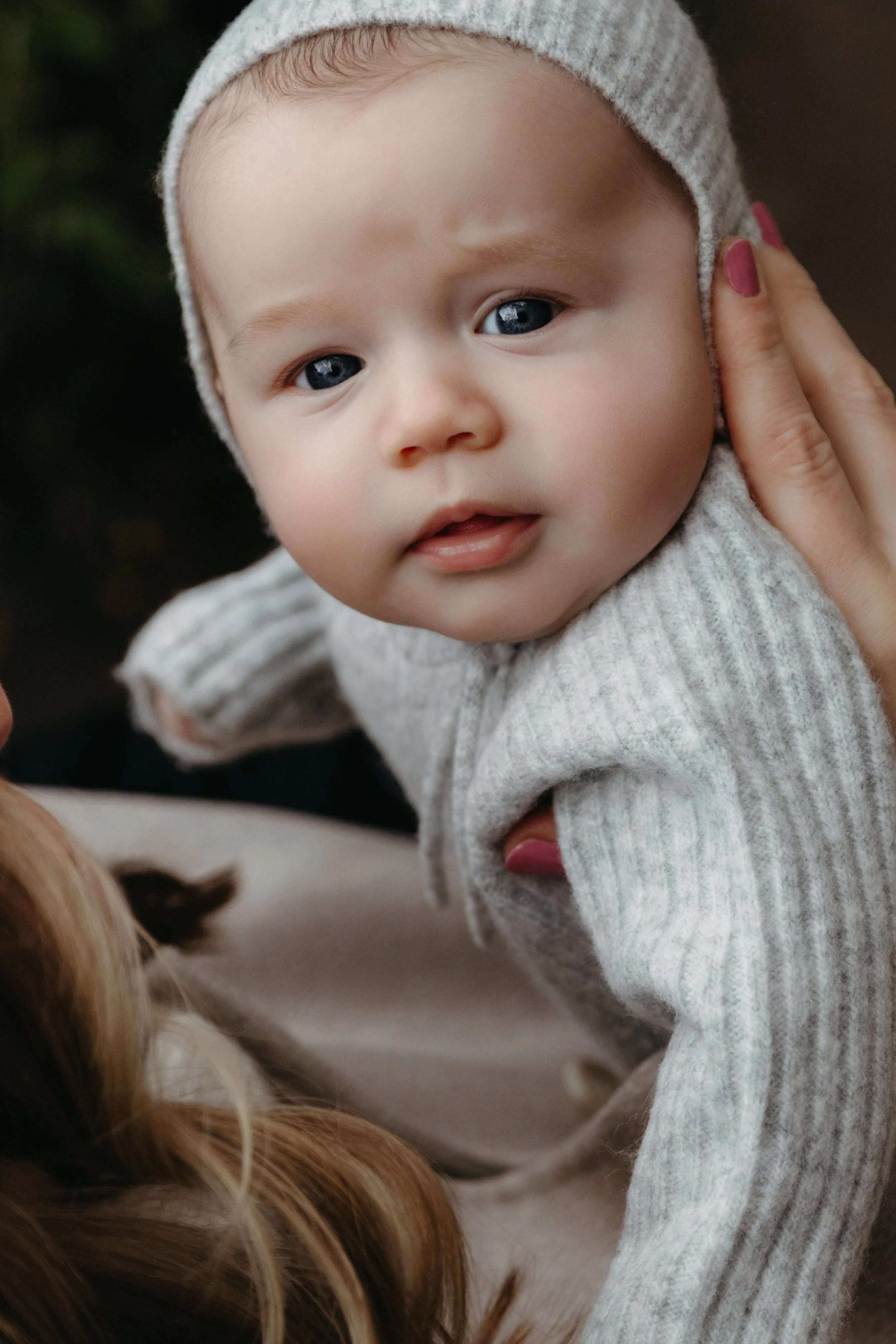 Close-up portrait of a newborn with blue eyes wearing a gray knitted hoodie, being gently held by an adult with pink-painted nails, with a blurred background. Photographed by Maison Mancel in Brooklyn.