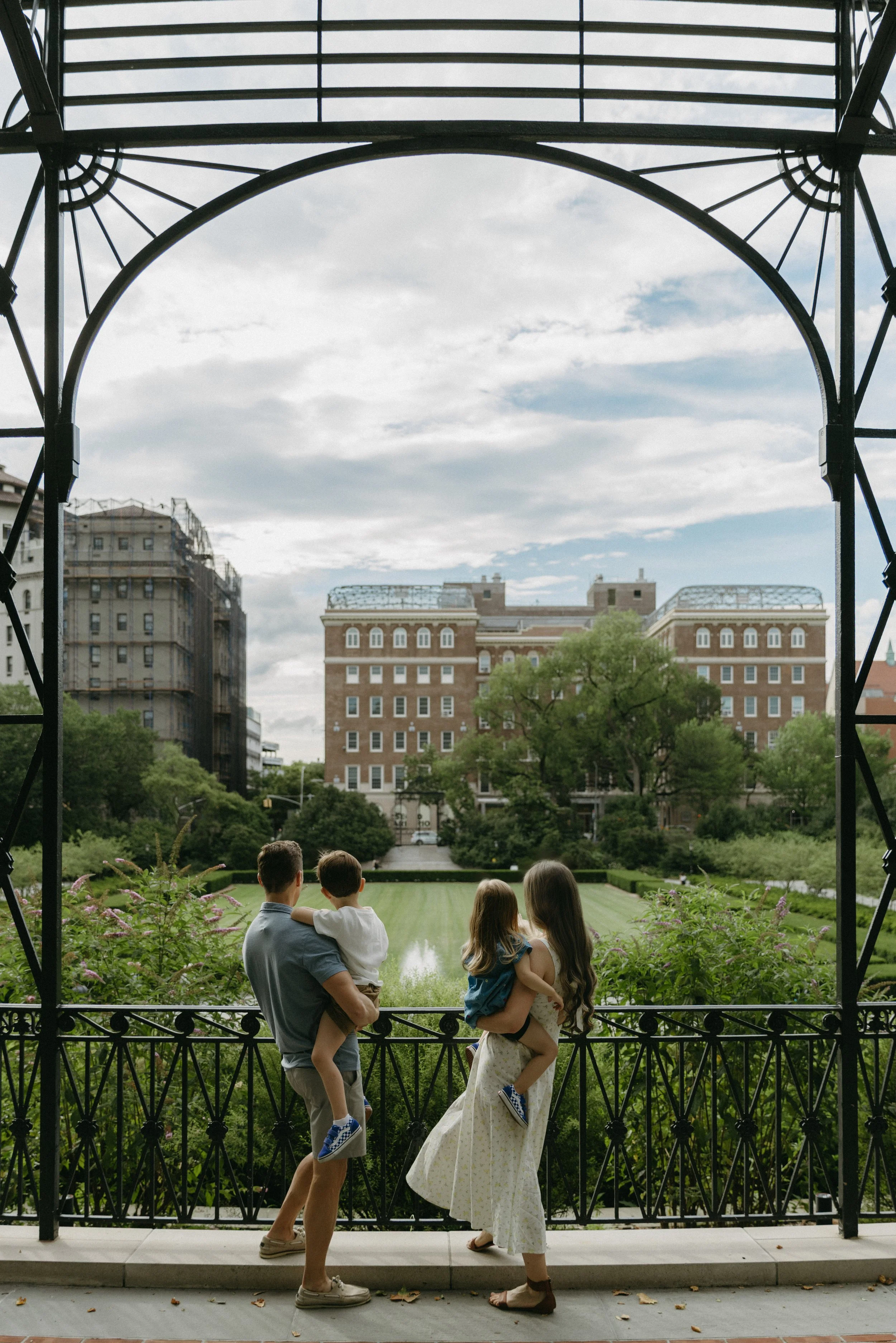 A family photograph of four with two children, a father and mother, standing on a balcony in Central Park, looking across a garden with a pond at a large brick building in the distance under a cloudy sky, by Maison Mancel.