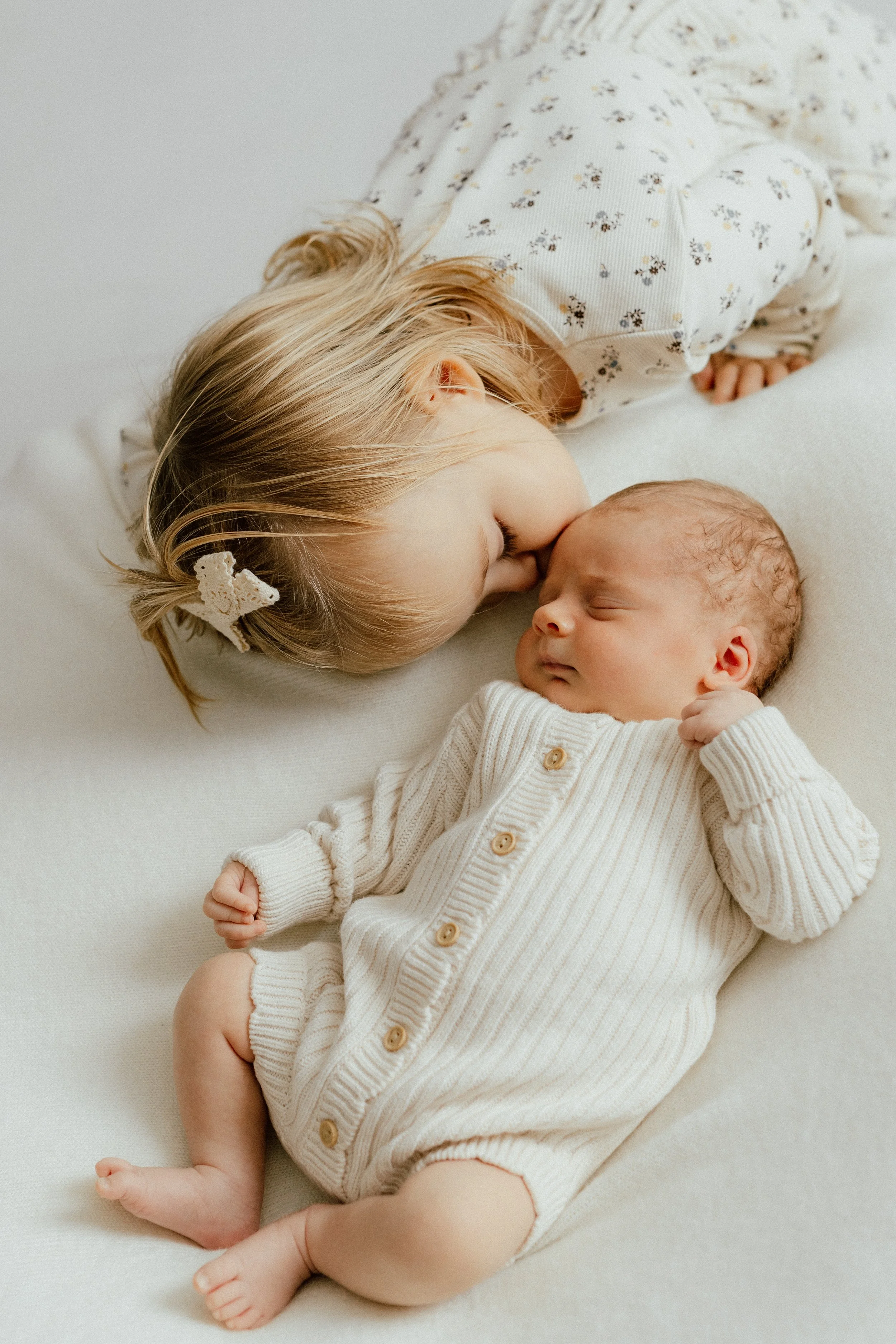 A newborn sibling portrait of a young girl and a newborn baby sleeping together on a white surface, with the girl resting her face on the baby's forehead. Photographed by Maison Mancel in NYC.