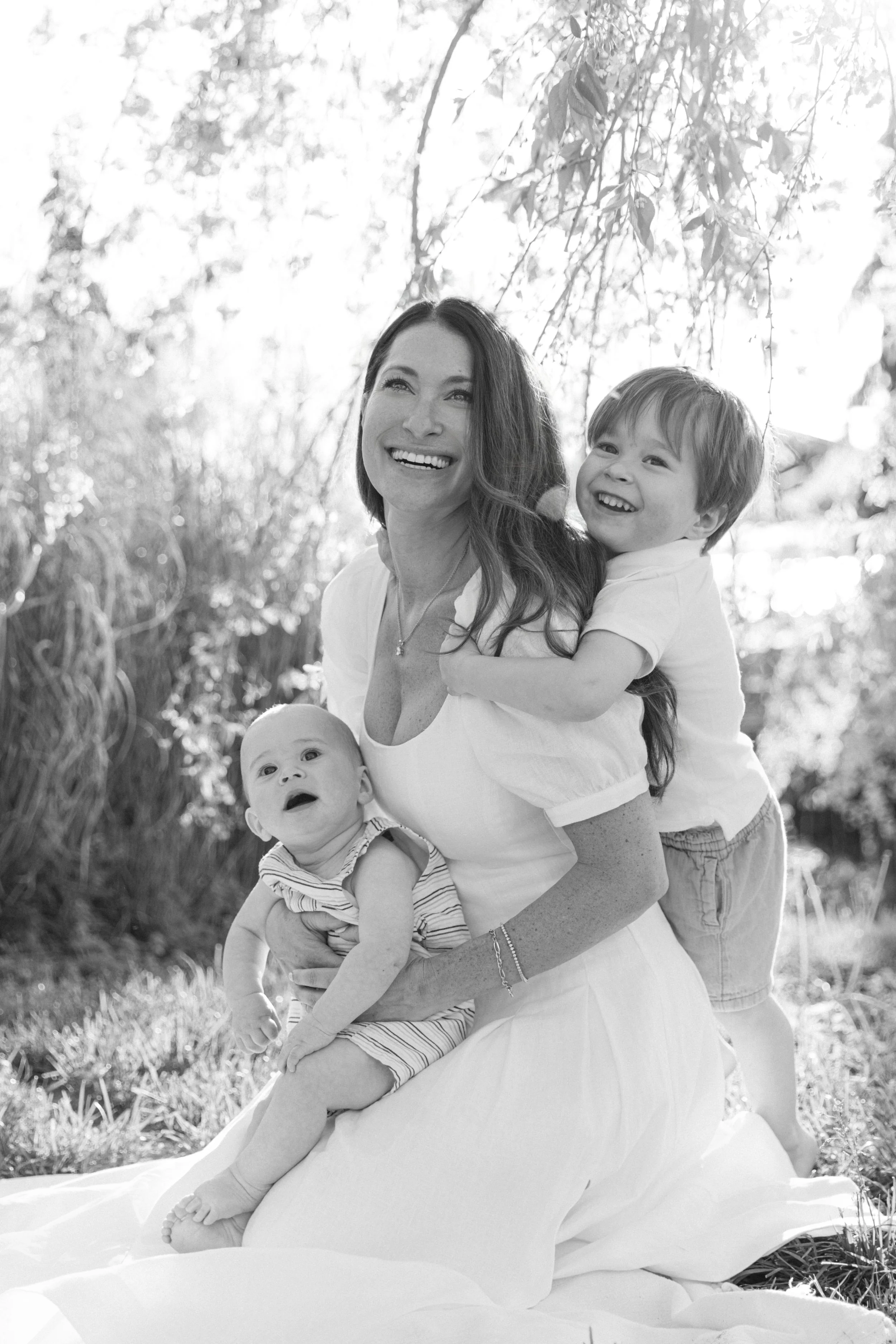 A smiling mother with two young children outdoors under trees in Brooklyn Heights, all in casual summer clothing, taken by Maison Mancel.