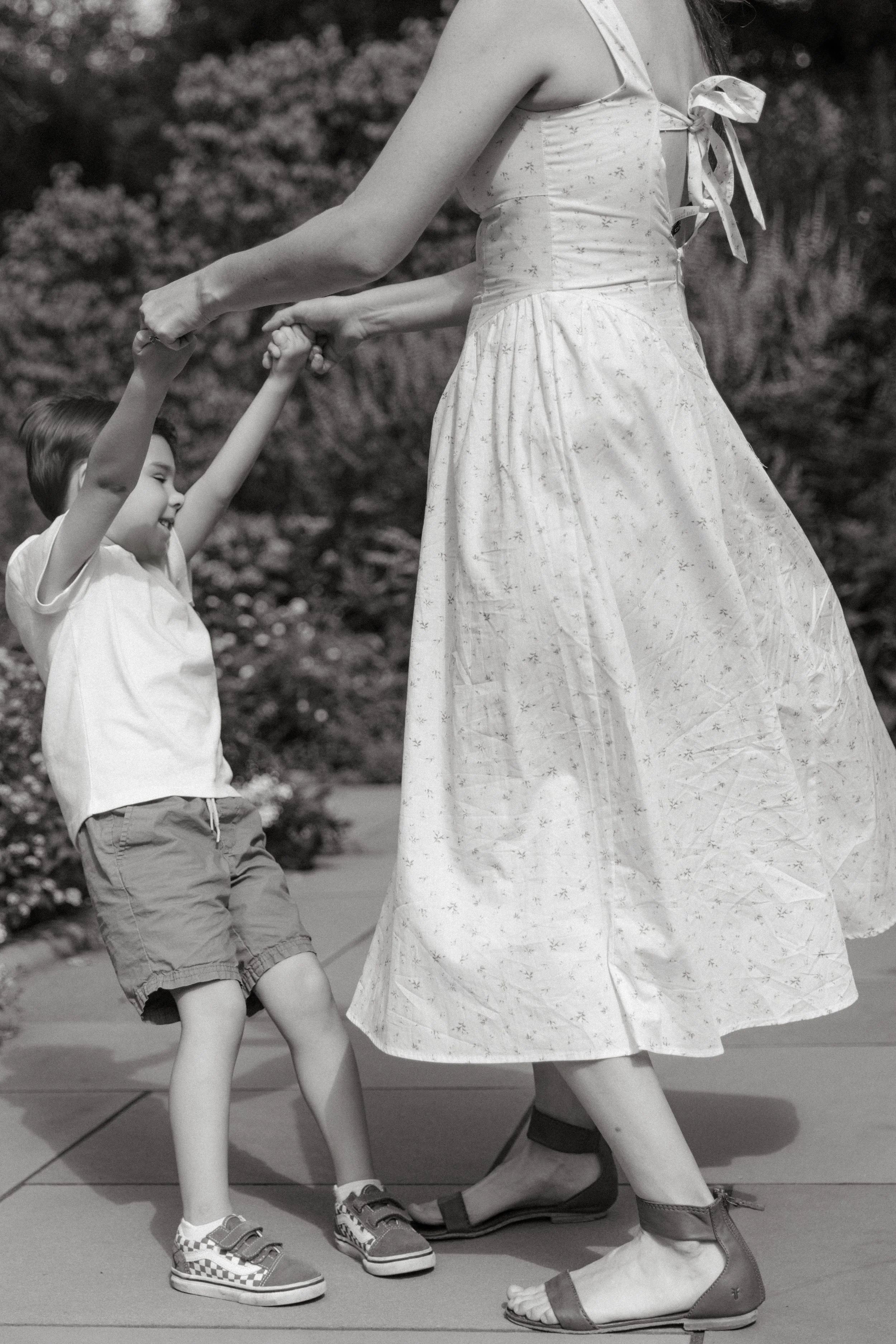 A family portrait of a mother holding a young boy's hands while they dance outdoors. The woman is wearing a light-colored dress, and the boy is dressed in a white shirt and shorts with sneakers, photographed by Maison Mancel in Central Park.
