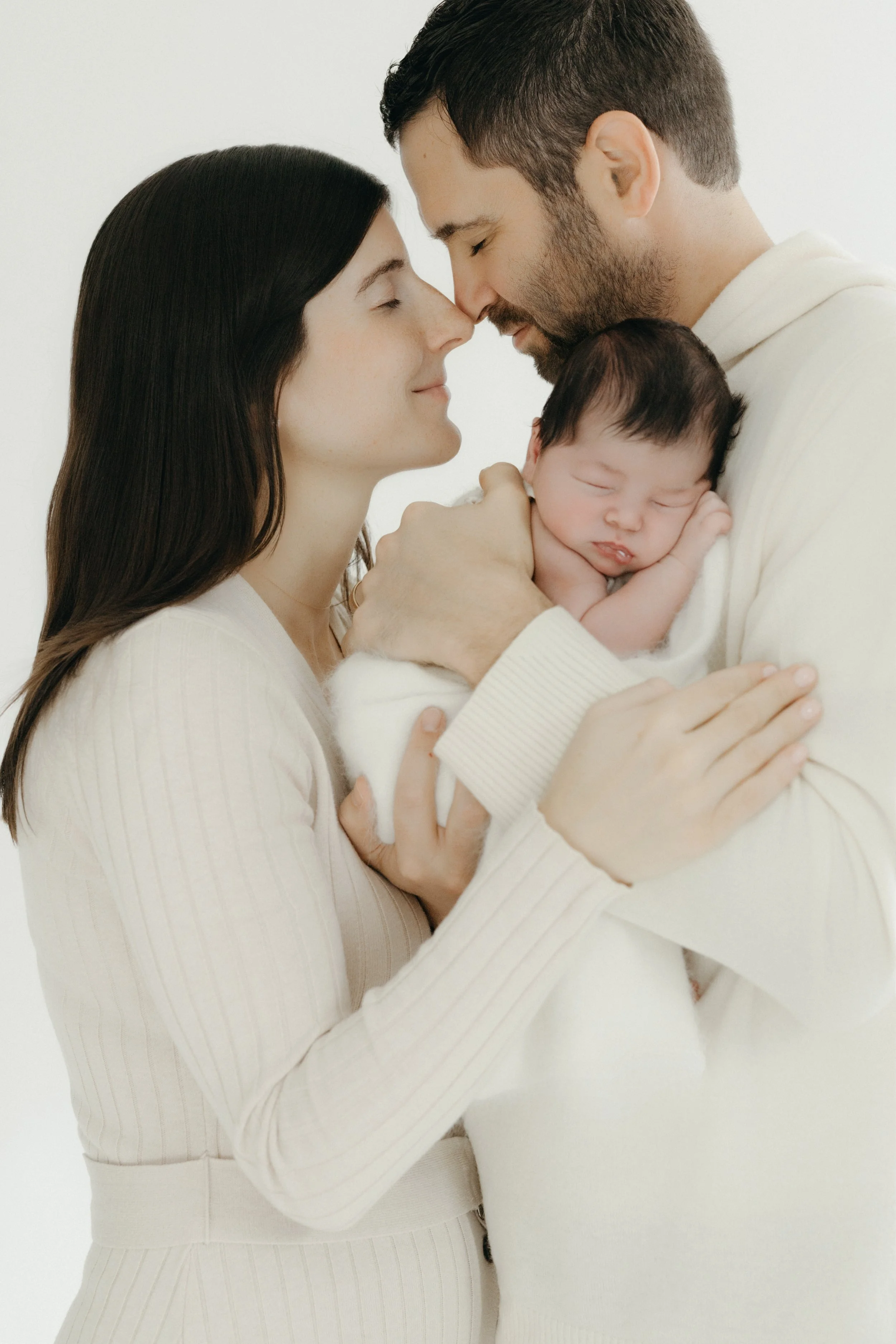 In-studio newborn portrait featuring young couple holding their sleeping newborn baby close, with foreheads touching, in a tender moment by Maison Mancel in NYC.