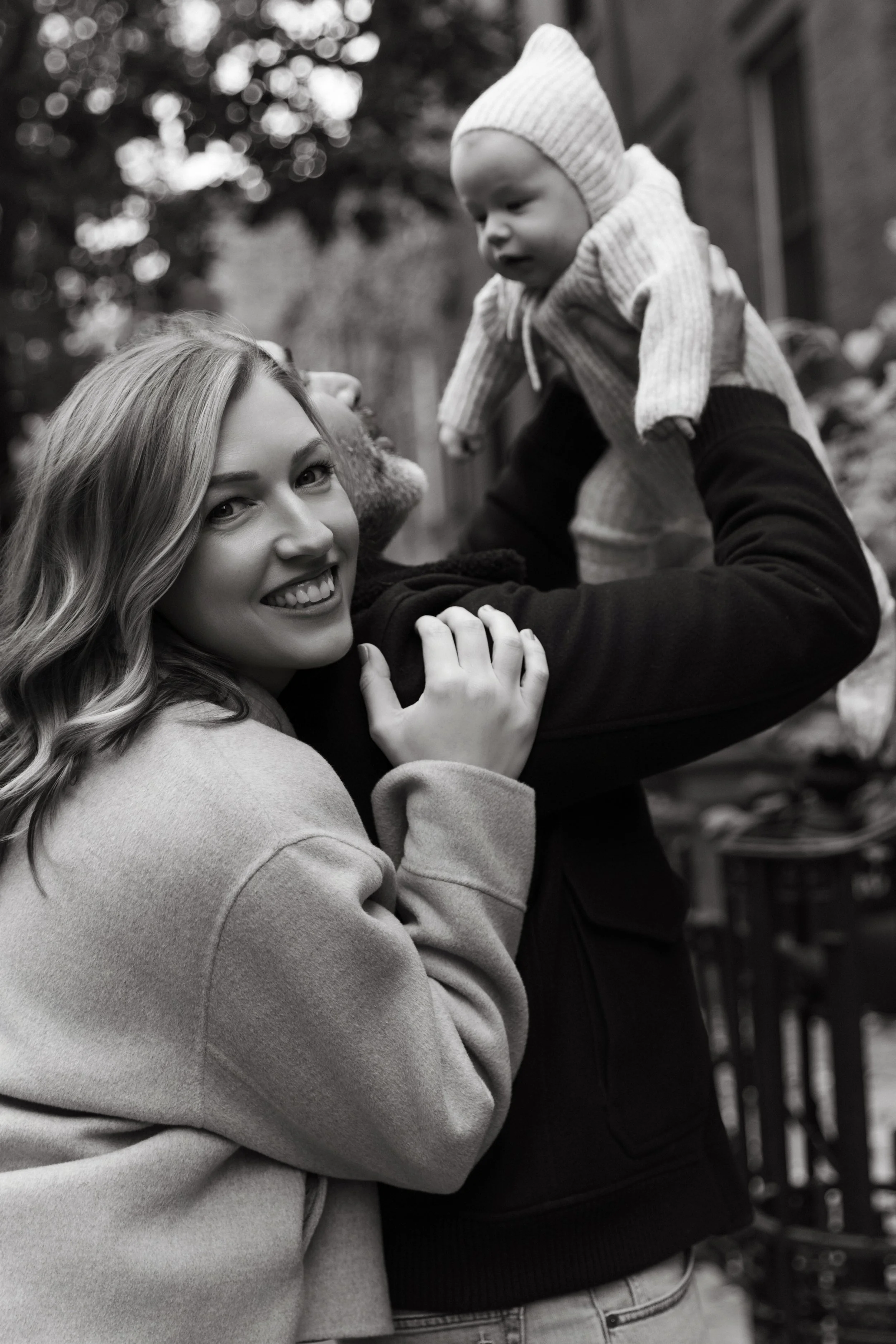 A black-and-white portrait of a mother smiling and holding a young child up in an outdoor setting with blurred trees and building in the background in Brooklyn Heights by Maison Mancel.