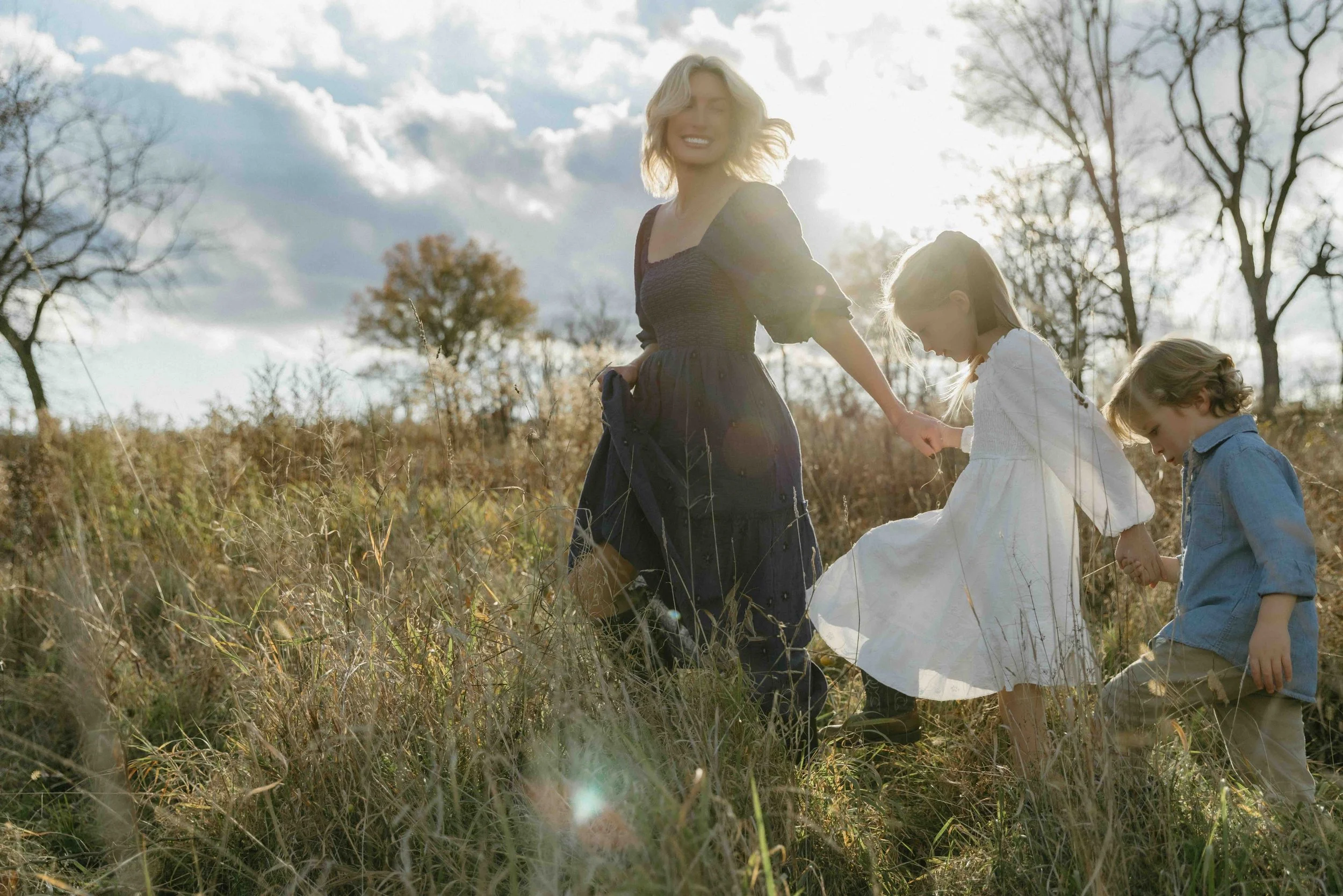 An outdoor family portrait of a mother and two children walking hand-in-hand through a grassy field during late afternoon with sunlight behind them, taken in New Jersey by Maison Mancel.