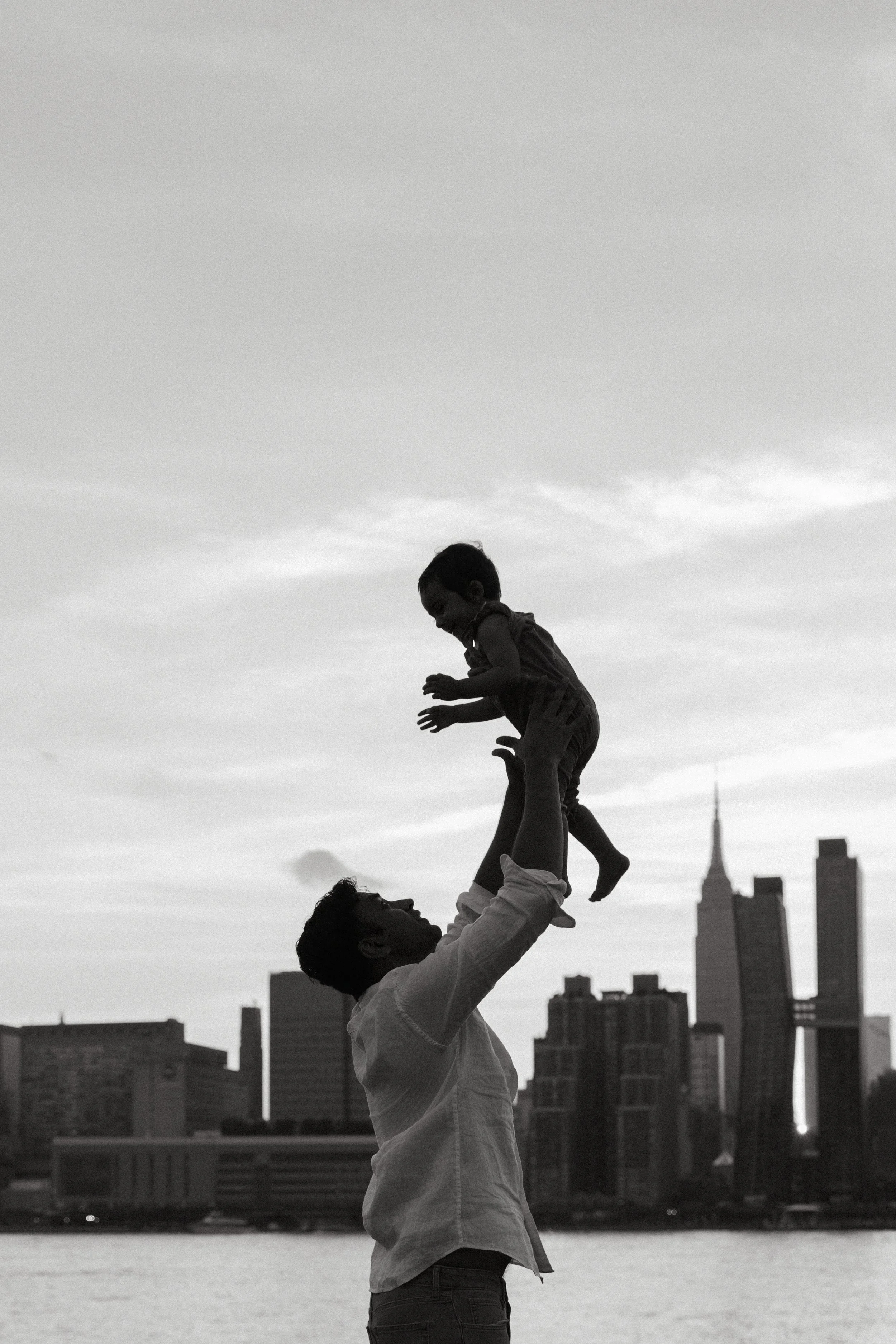 A family portrait featuring a father lifting a child above their head with a New York City skyline in the background, in black and white, by Maison Mancel.