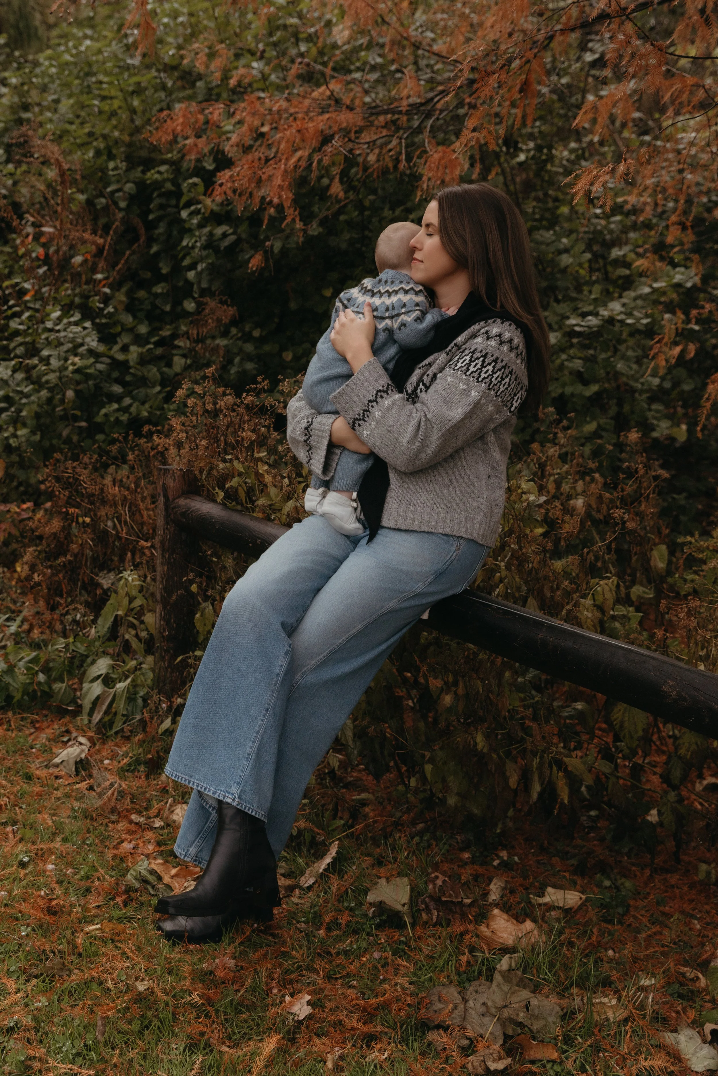 A family portrait of a mother sitting on a wooden fence holding a baby while surrounded by autumn foliage in Brooklyn Bridge Park, by Maison Mancel.