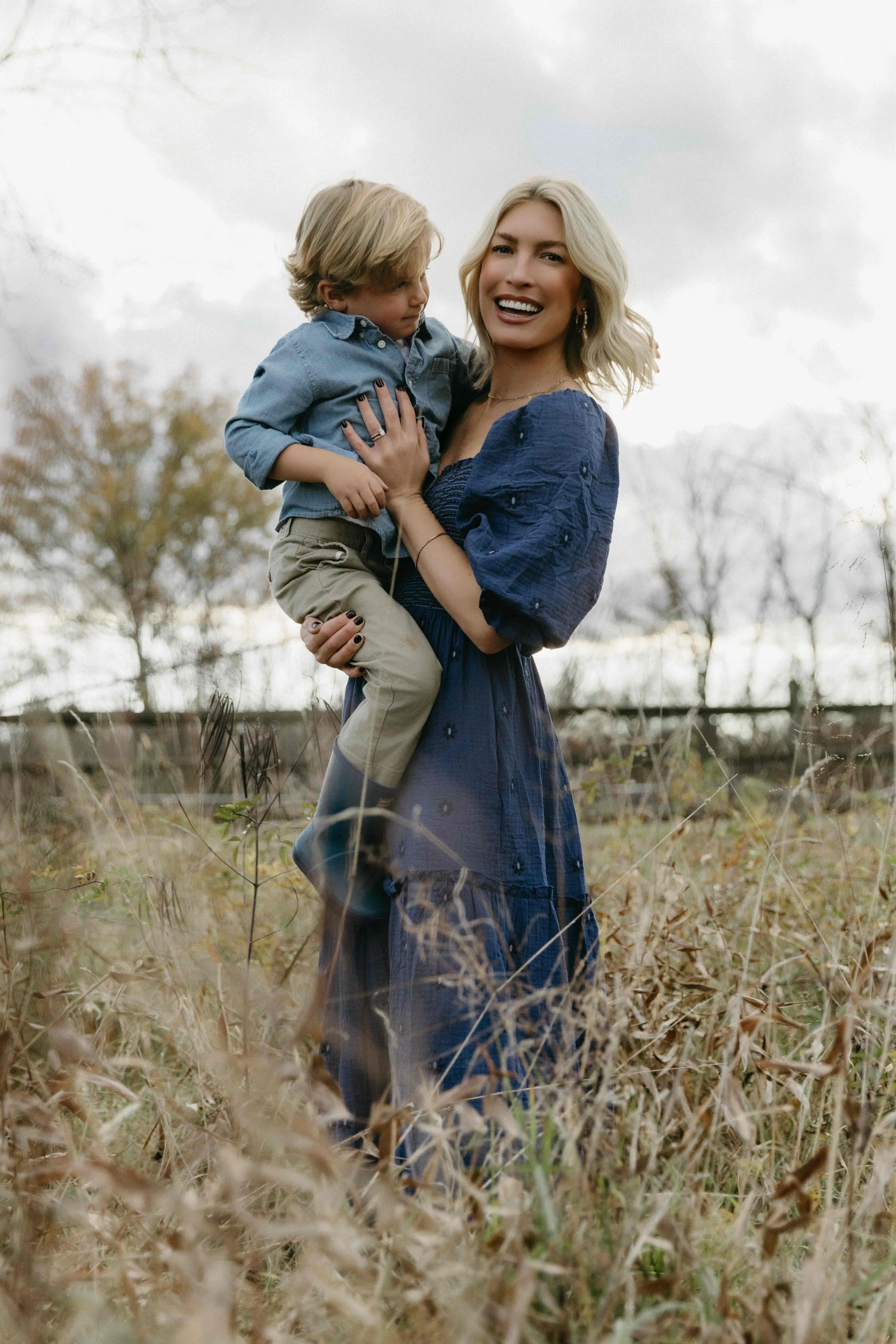 NYC family photograph of a mother holding a young boy outdoors in a field with trees and overcast sky in the background - Maison Mancel.