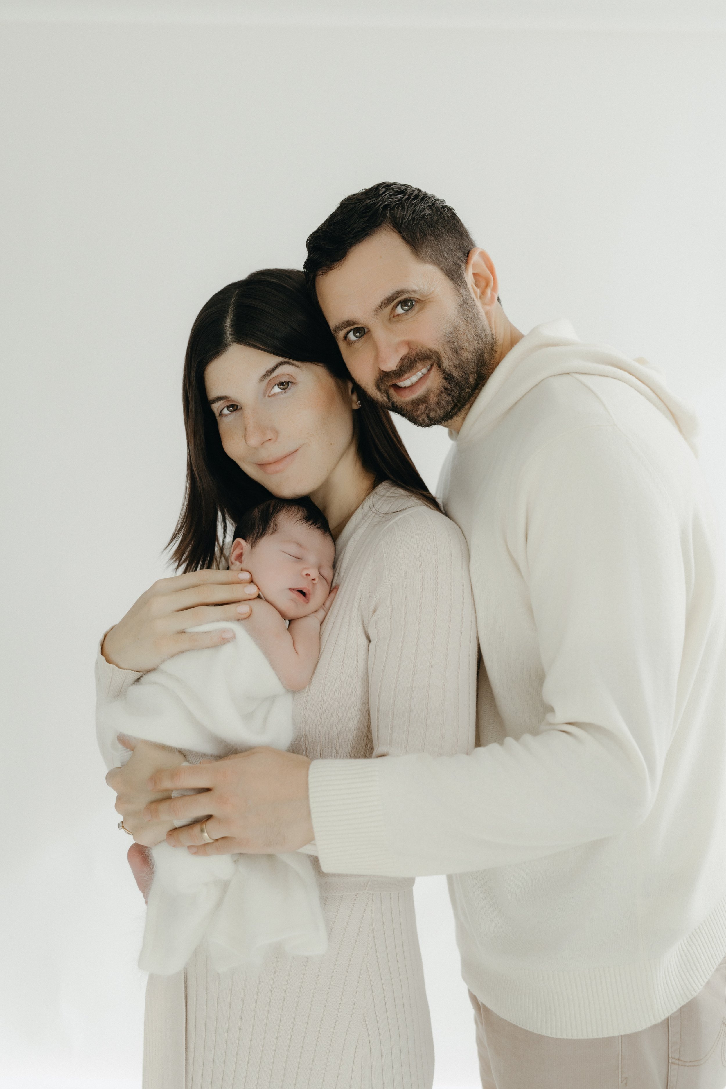 A happy family of three, a woman holding a sleeping baby, with a man beside her smiling in a bright, white background. Photographed by Maison Mancel in NYC studio.