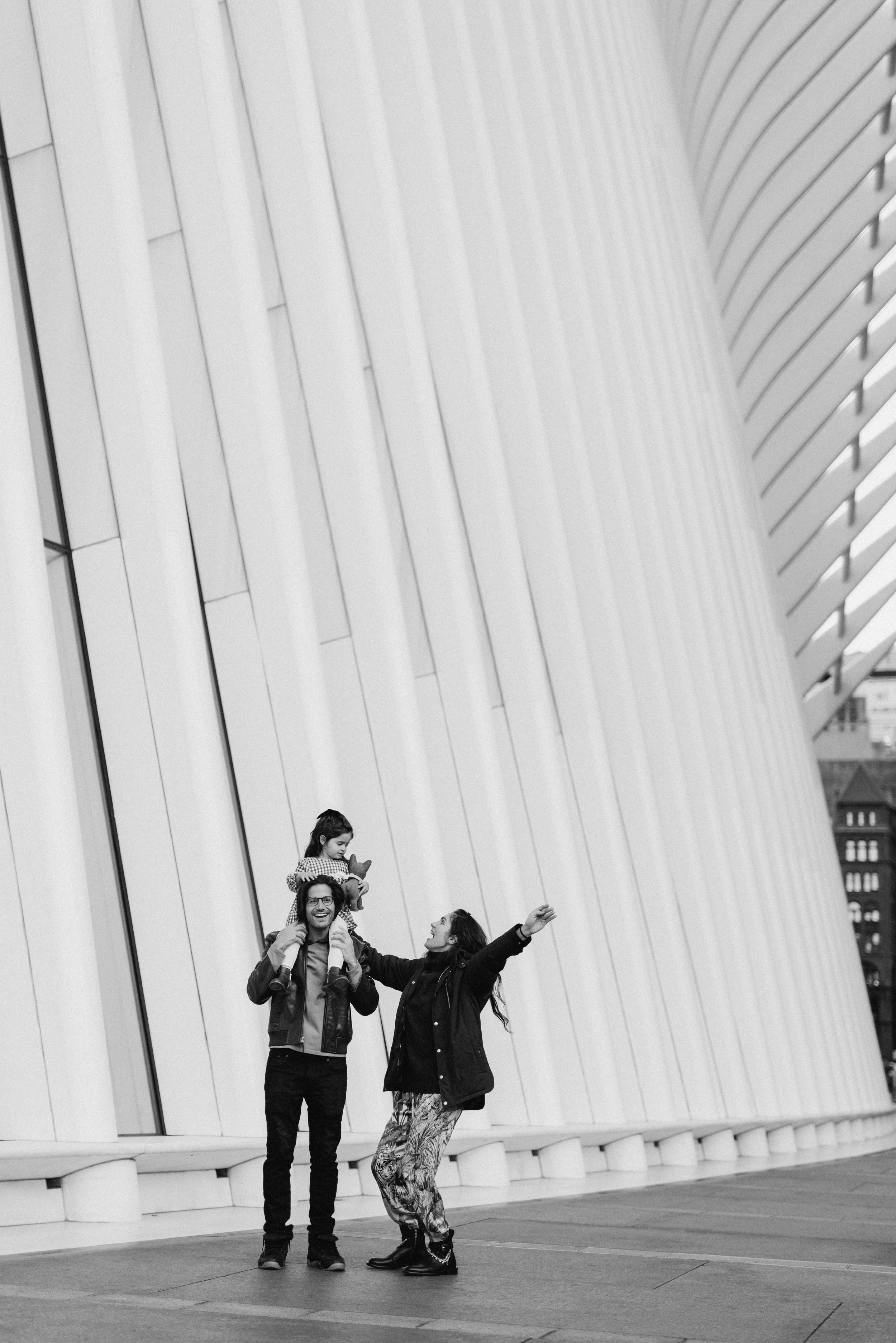 A family of three enjoying time together outside a modern building with curved white panels. The father carries the daughter on his shoulders, while the mother gestures happily toward them. Photographed by Maison Mancel in lower Manhattan.