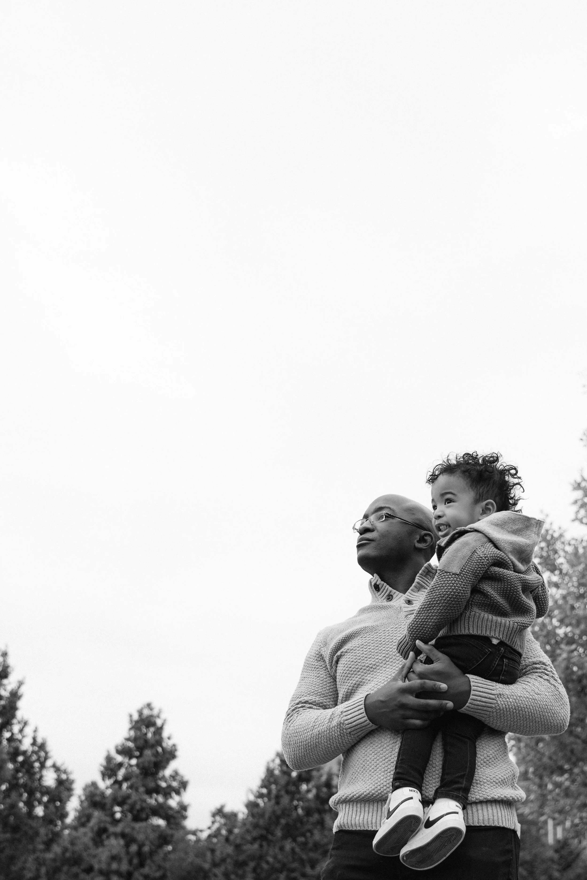 A family portrait of a father holding a young child in an outdoor park in Brooklyn with trees, both looking up into the sky, by Maison Mancel.