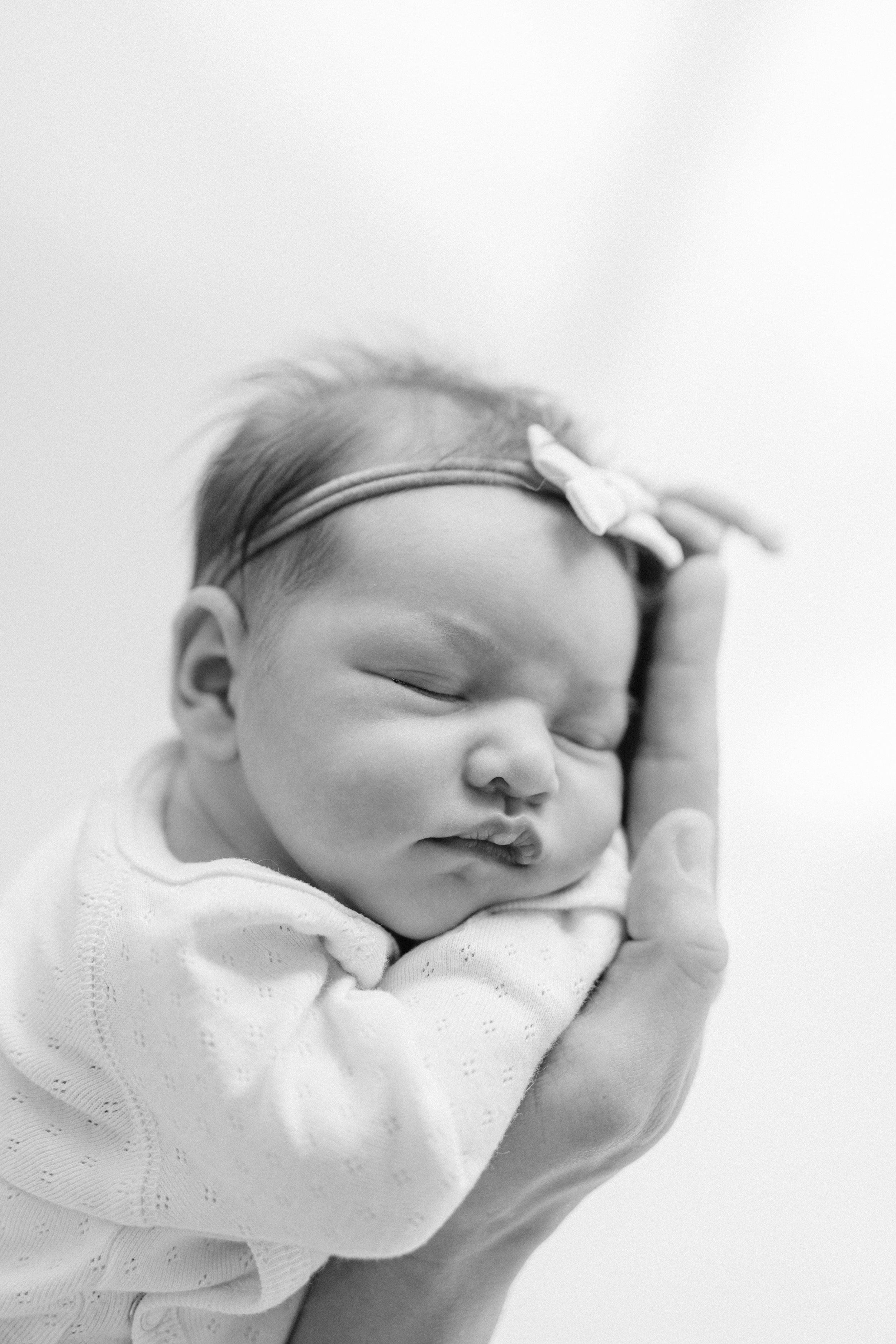 Black and white photo of a sleeping baby, gently held by a hand with the baby's head resting on their palm, wearing a headband with a small bow. Photographed in-studio by Maison Mancel in NYC.