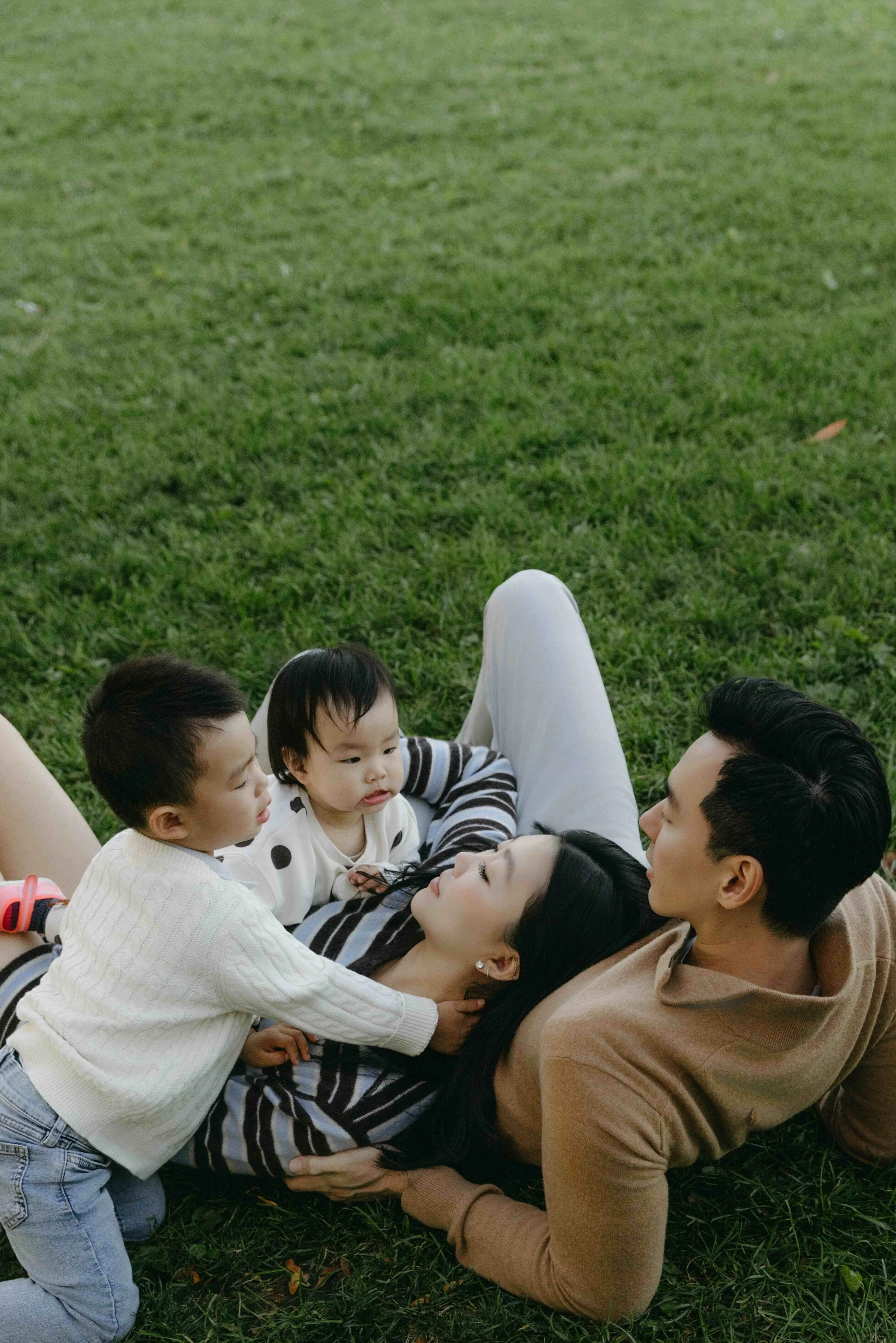 Family portrait showing a family of four lying on grass in Brooklyn with children interacting with their mother and father in an outdoor setting - Maison Mancel.