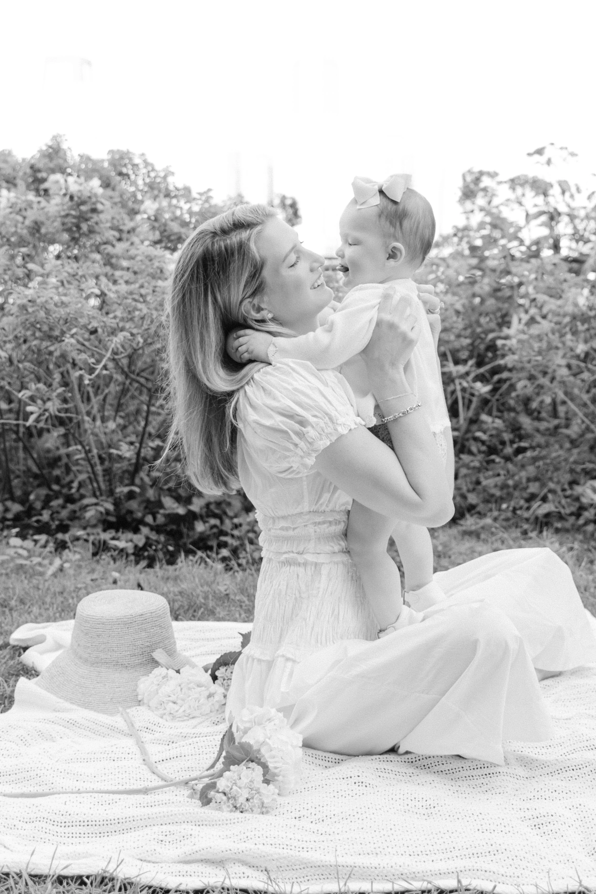 A photograph of a mother sitting on a blanket holding a young girl in an outdoor setting, both smiling and looking at each other, with a hat and flowers on the blanket, photographed by Maison Mancel in Battery Park City.