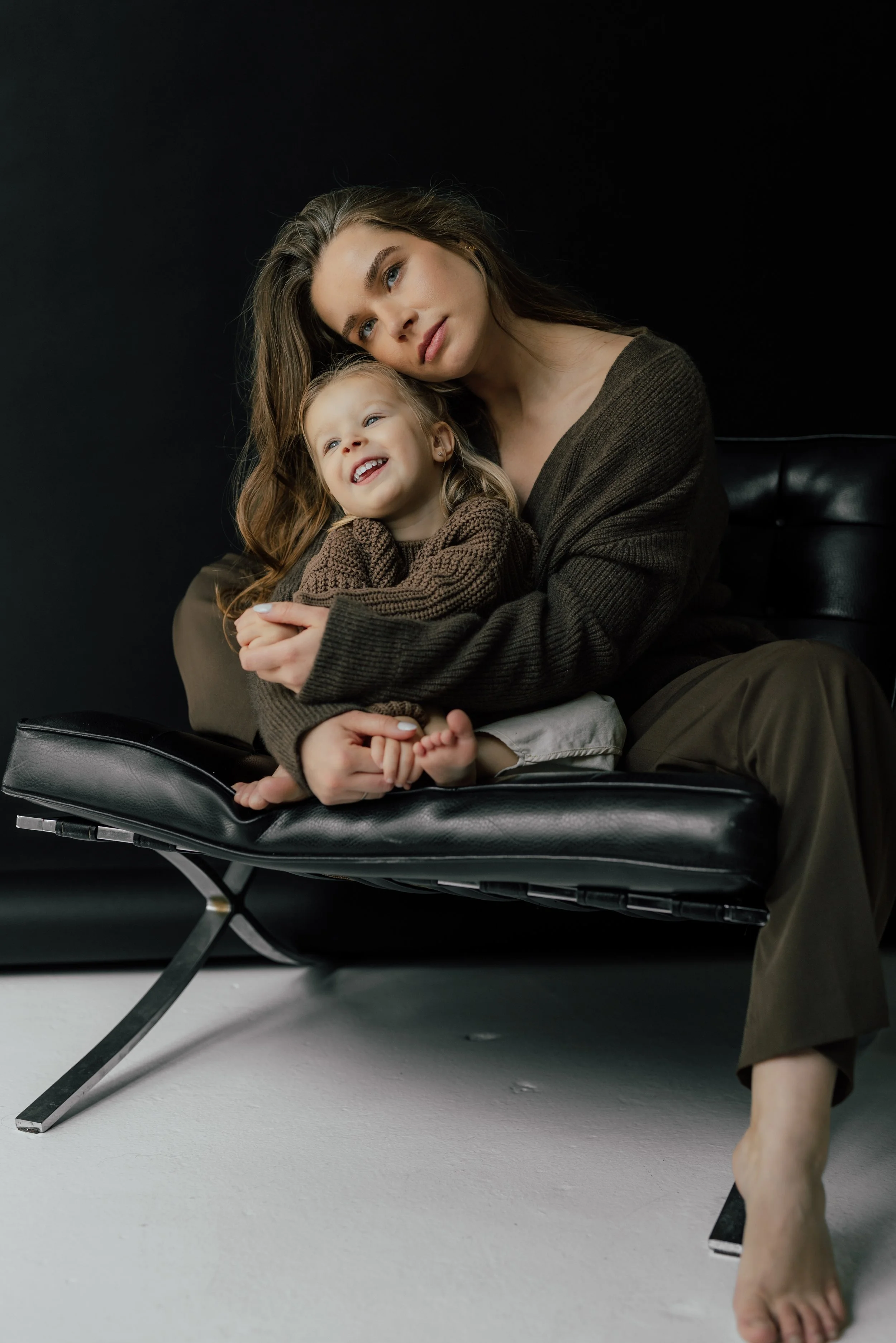 A studio portrait of a mother and a young girl sitting on a black leather chair with a dark background; the mother is hugging the girl, who is smiling, and they are both looking slightly to the left, photographed by Maison Mancel.