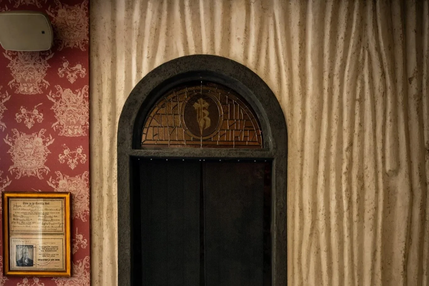 Elevator with decorative stained glass window featuring a Chinese character, set into an ornate wall with textured curtains on one side and red damask wallpaper with a framed document and photograph on the other.