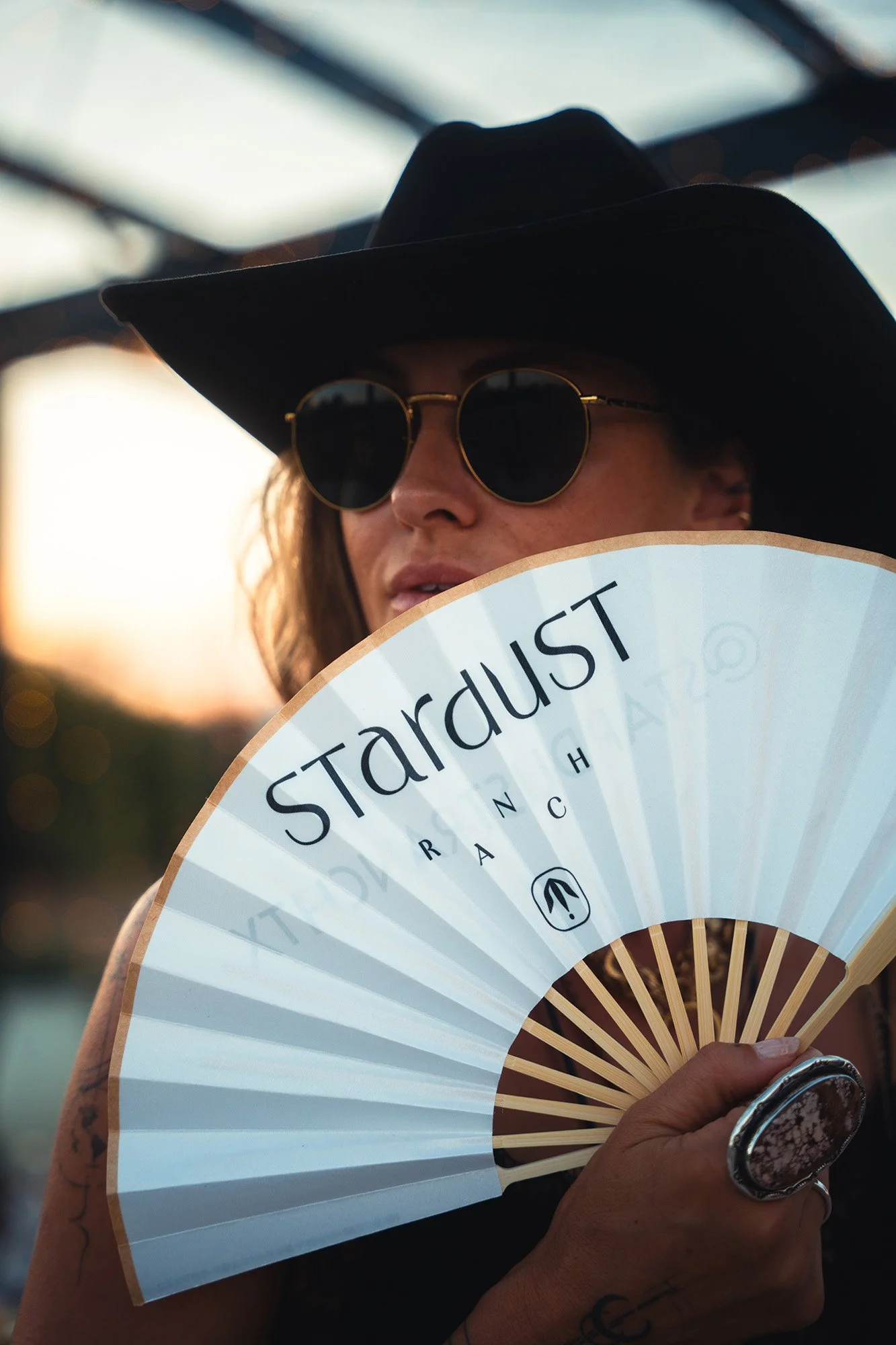 A person wearing sunglasses and a black wide-brimmed hat holding a fan with the word 'August' written on it, partially covering their face.