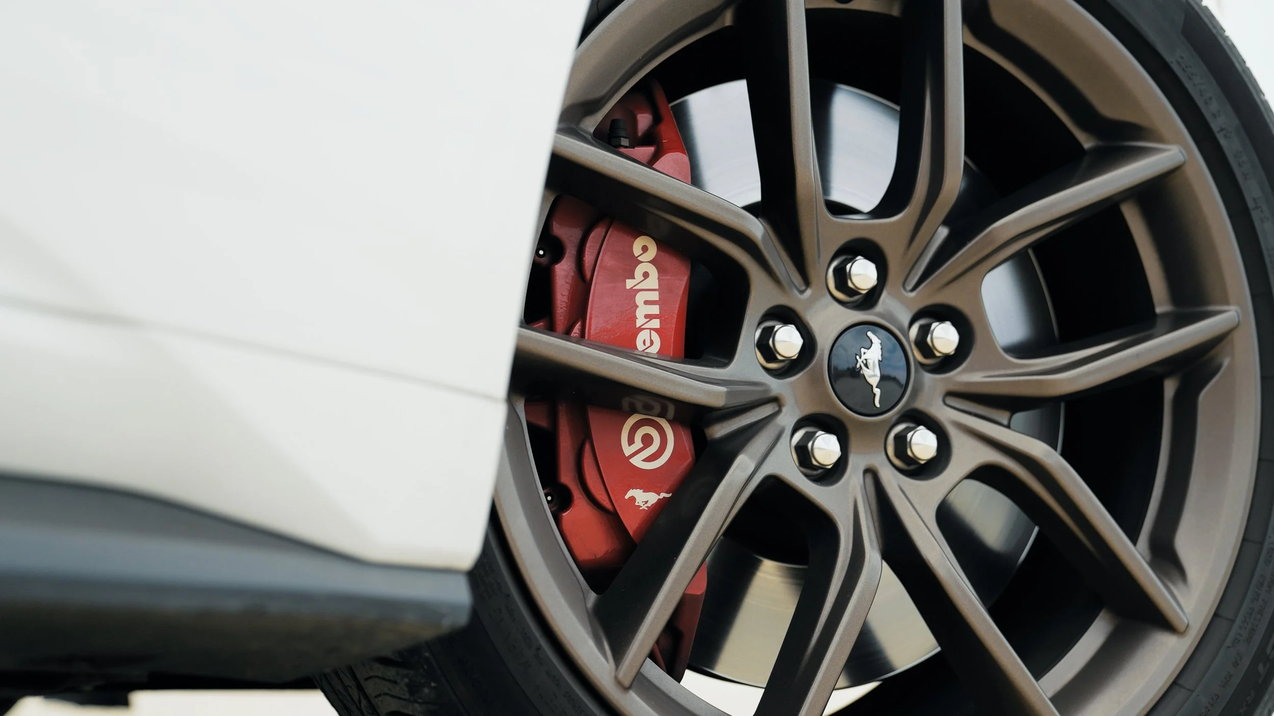 Close-up of a car wheel with a Mustang logo, red Brembo brake caliper with Brand logo, and multi-spoke alloy rim.