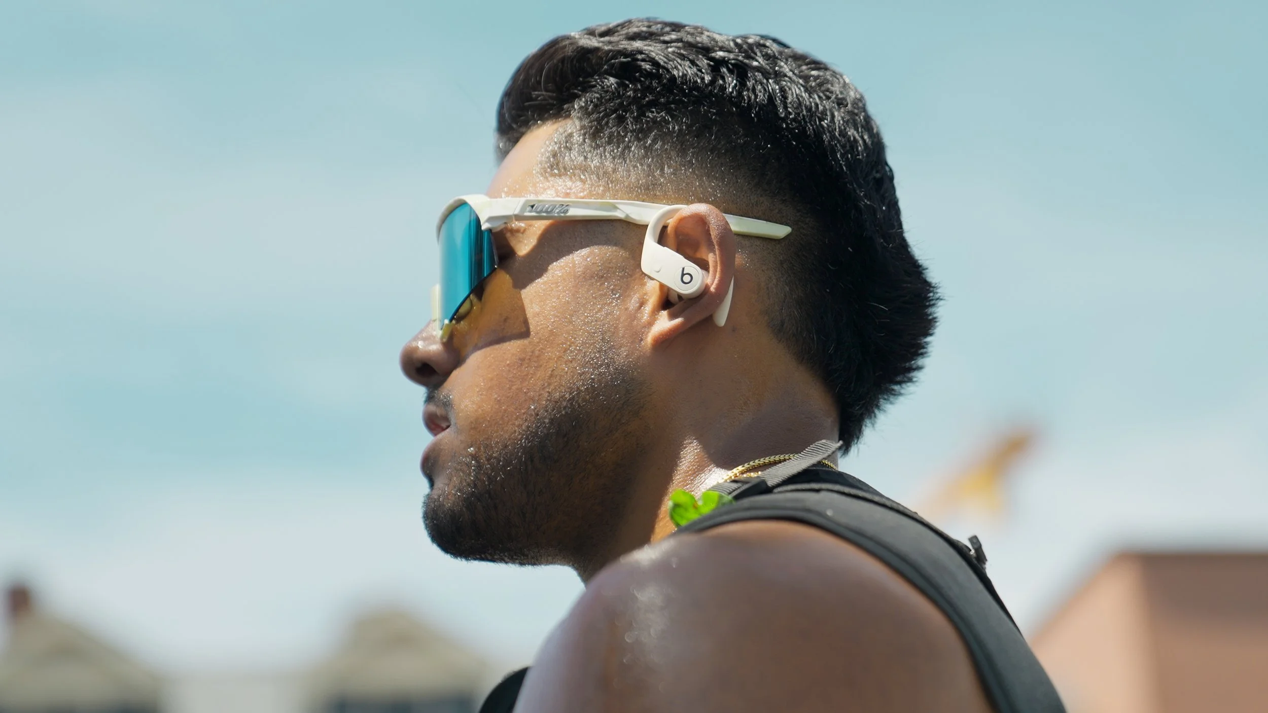 Close-up of a man with wet hair wearing sunglasses and earbuds, outdoors in bright sunlight.