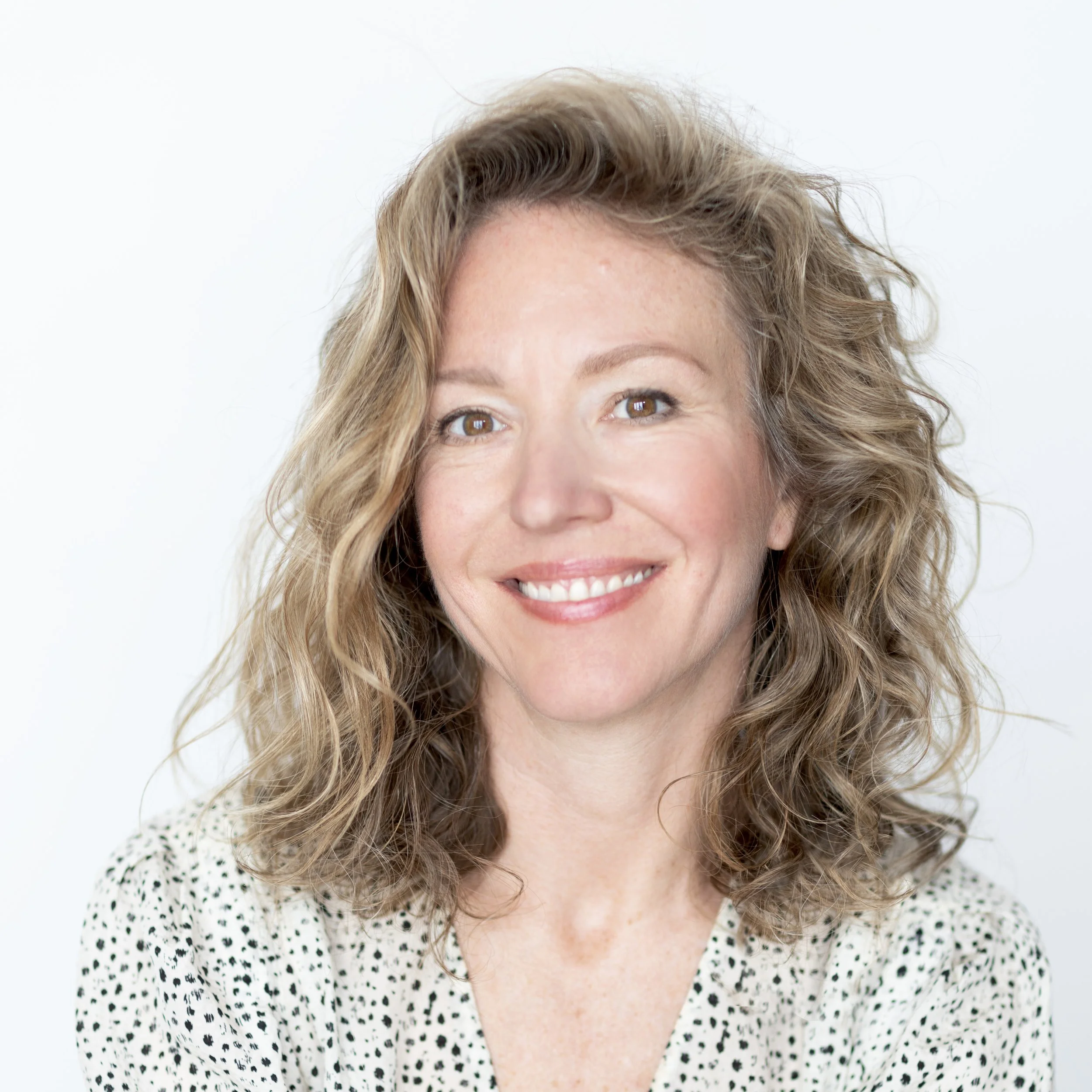 A person with wavy blonde hair, smiling, wearing a light-colored patterned blouse, against a plain background.