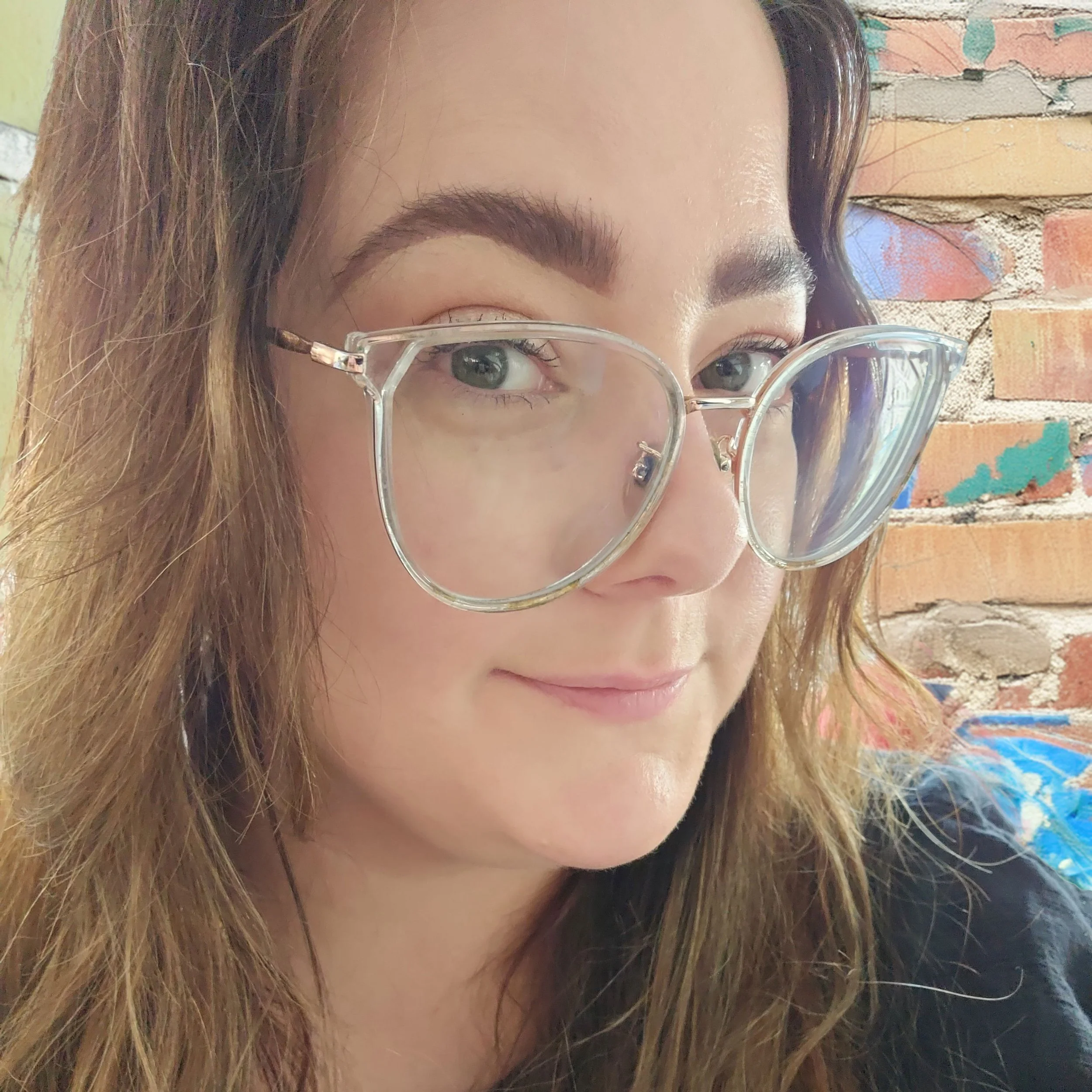 Close-up of a woman wearing large transparent glasses with a brick wall background.