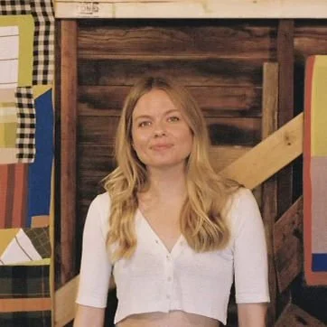 A woman with long blonde hair wearing a white top, standing in front of a wooden wall decorated with colorful quilts. The setting appears rustic with wooden elements and patchwork fabric.