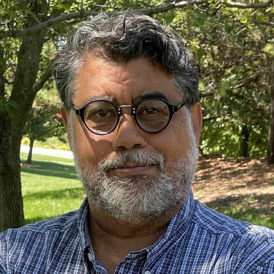 Man with glasses and facial hair standing outdoors near trees and grass.