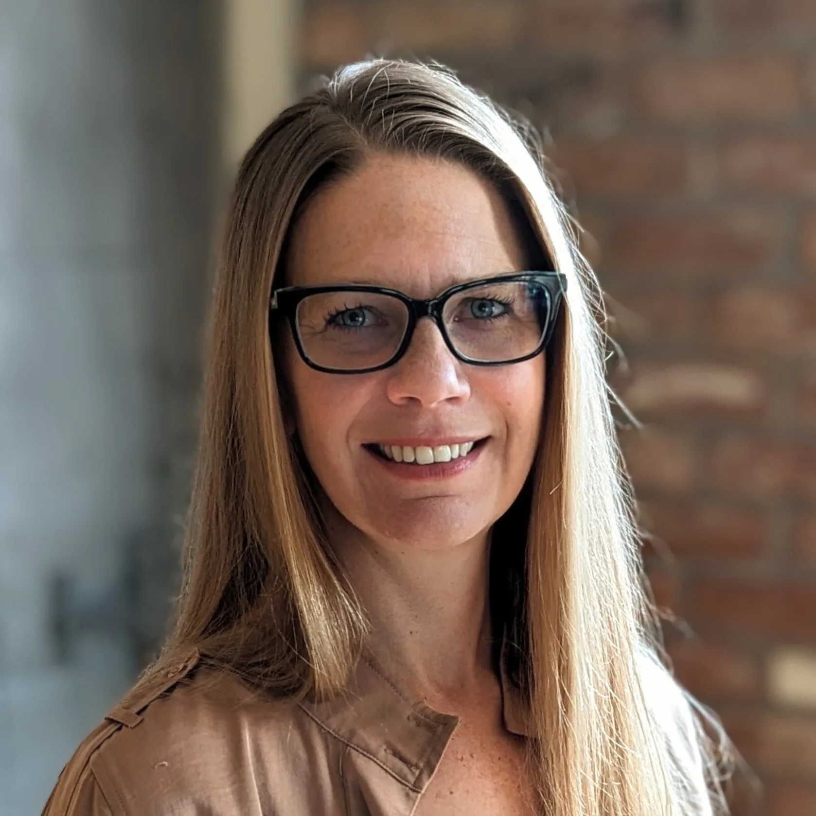 Person with long hair and glasses smiling in front of a brick wall.