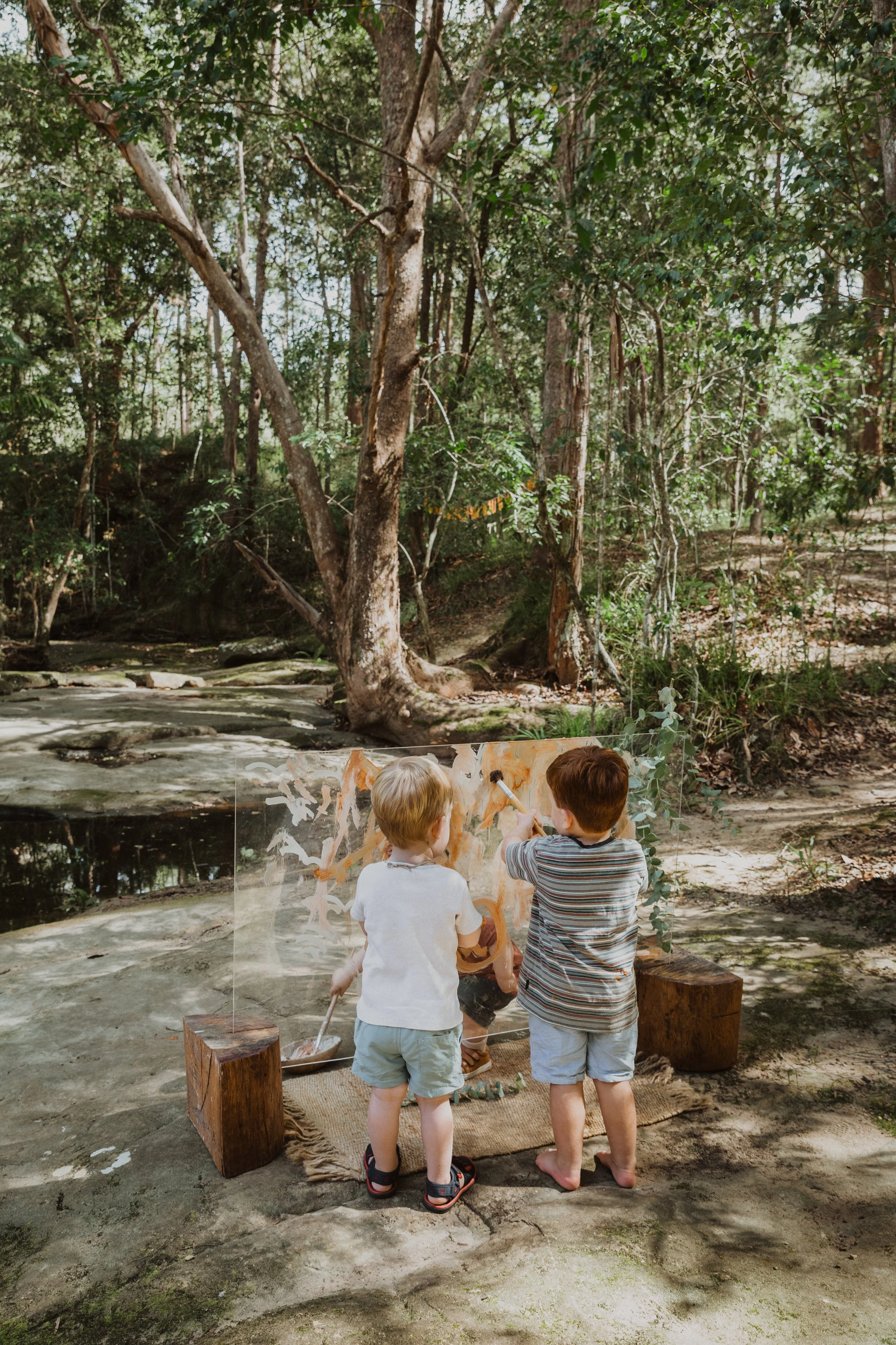 Two young boys are painting a large canvas outdoors in a wooded area, sitting on a woven rug with wooden blocks around them, with a backdrop of trees and a stream.