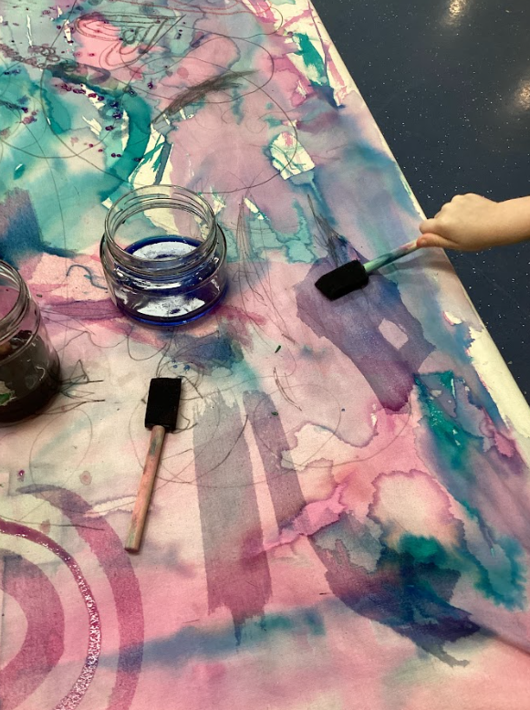 Child using a foam brush to paint on a colorful, watercolor-style art canvas with jars of paint nearby.